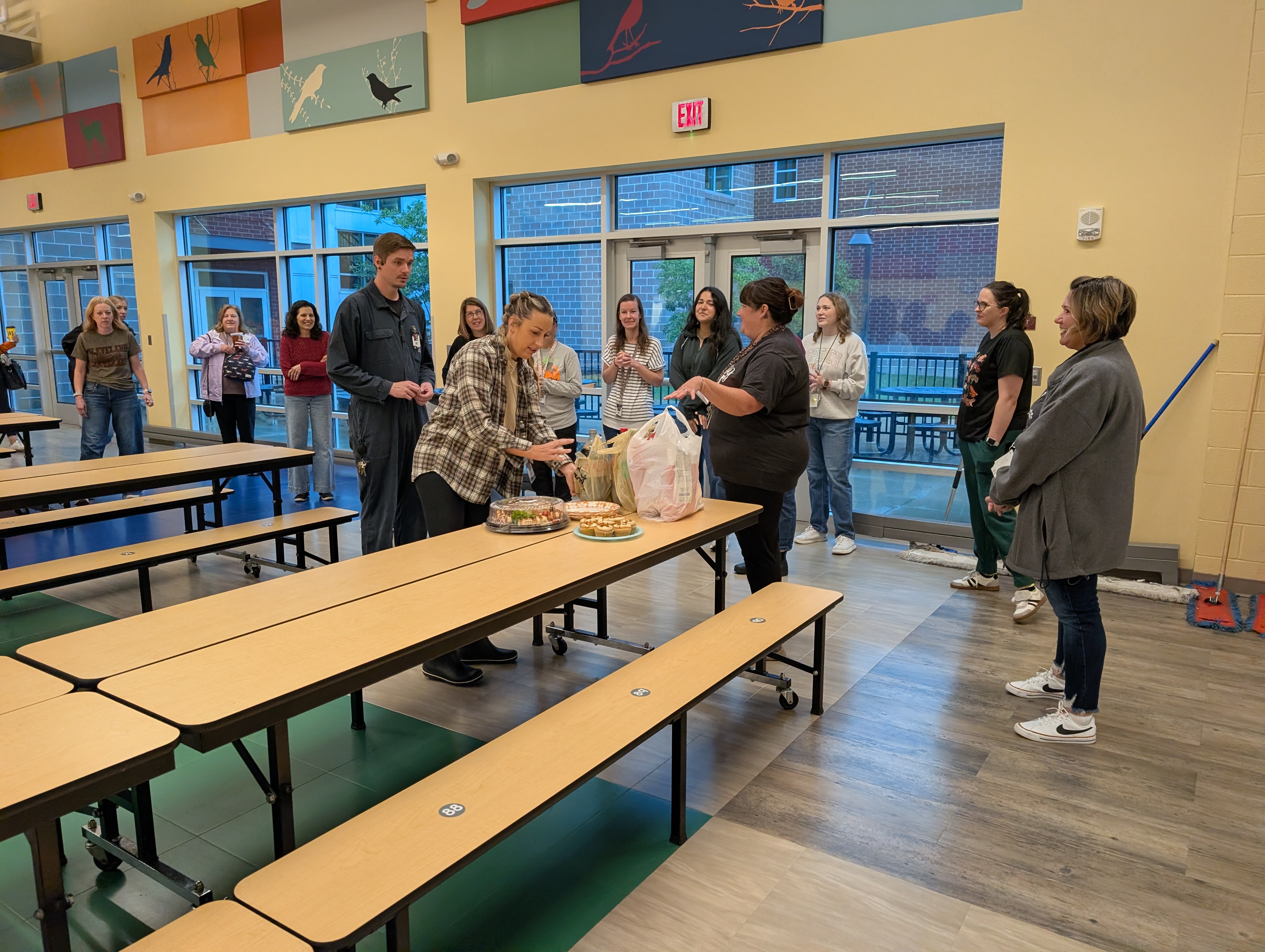 A group of adults gathers in a bright cafeteria with yellow walls, setting up food on a long table.