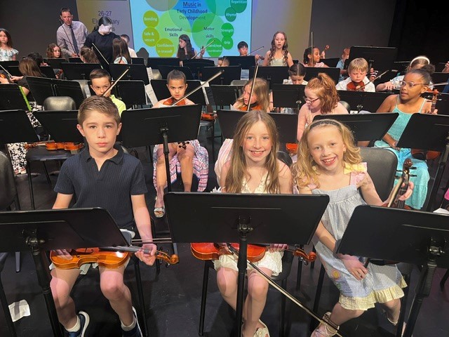 Children in an orchestra sit with violins and music stands, ready to play.