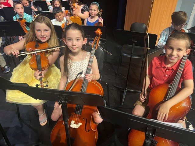 Three smiling children sit with their string instruments in an orchestra setting.