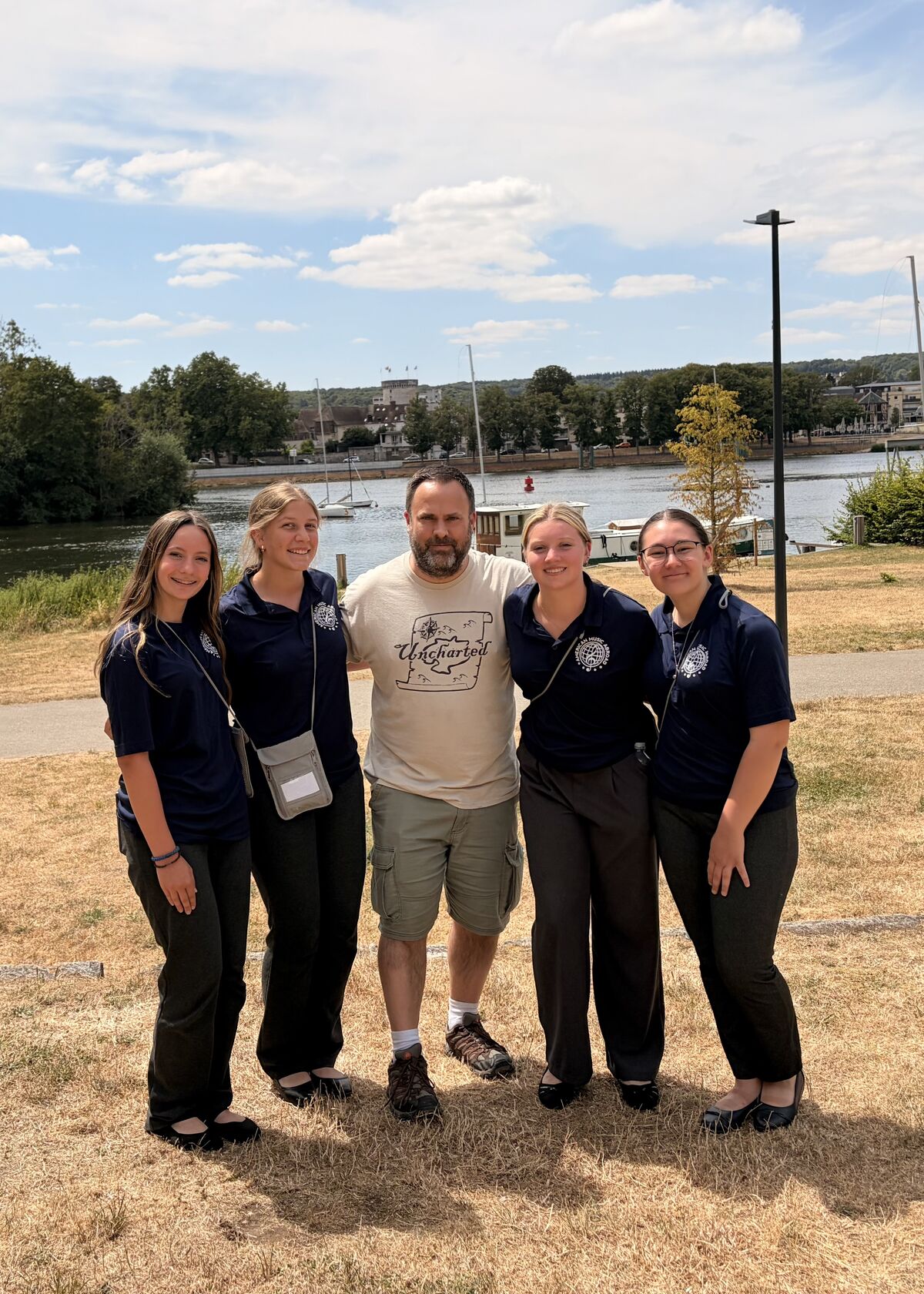 A man in a beige t-shirt stands with four young women in navy blue shirts, posing outdoors by a river.