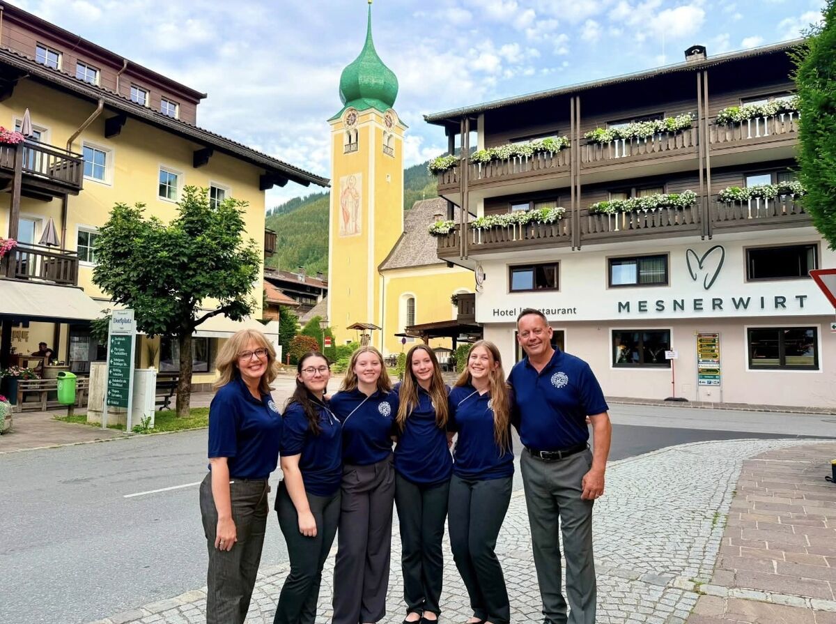 A group of six people, two adults and four young women, stand together in front of a hotel and restaurant called Mesnerwirt, with a church tower in the background.