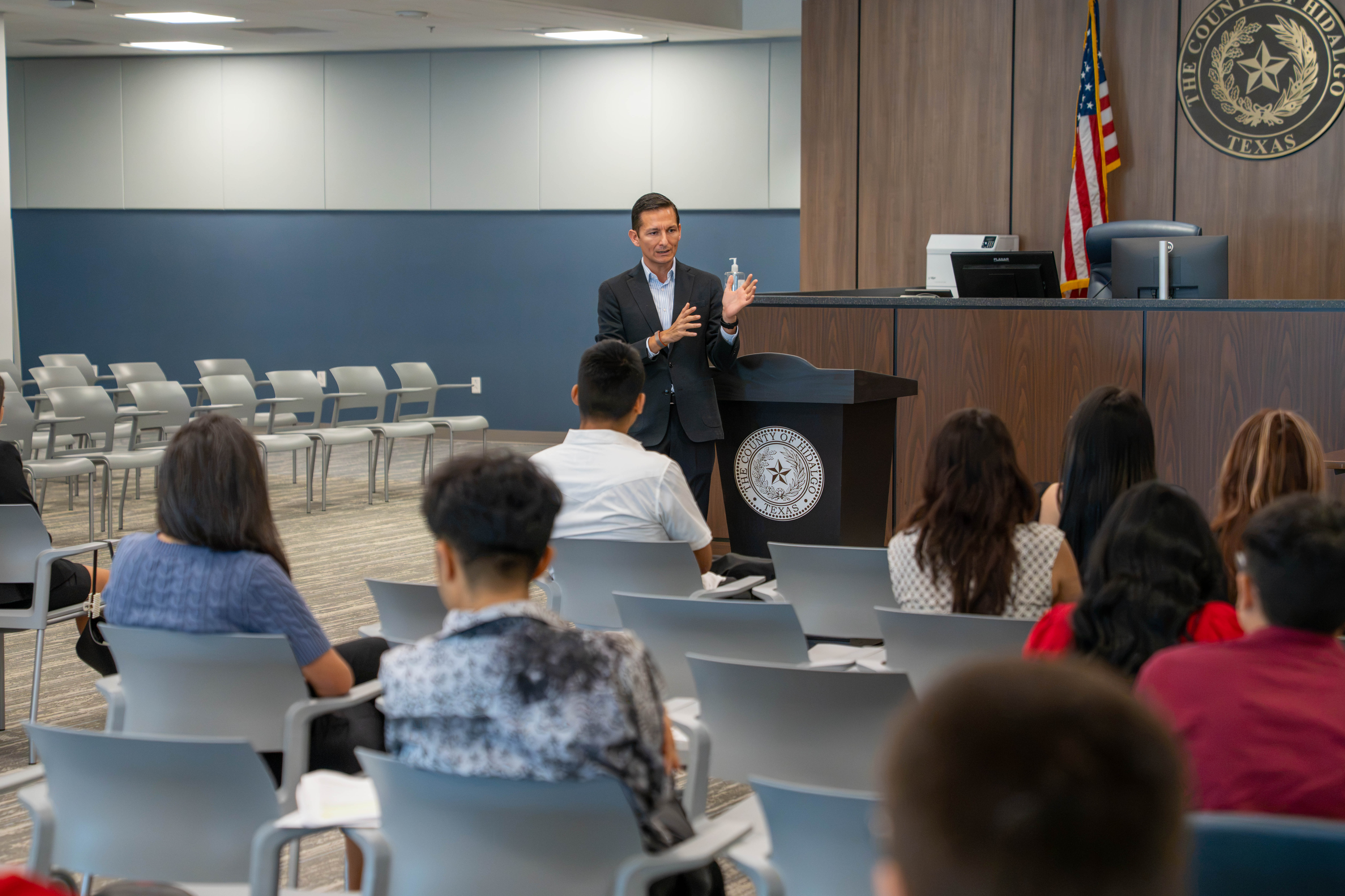 A man in a suit gestures while speaking to an audience seated in a modern conference room.