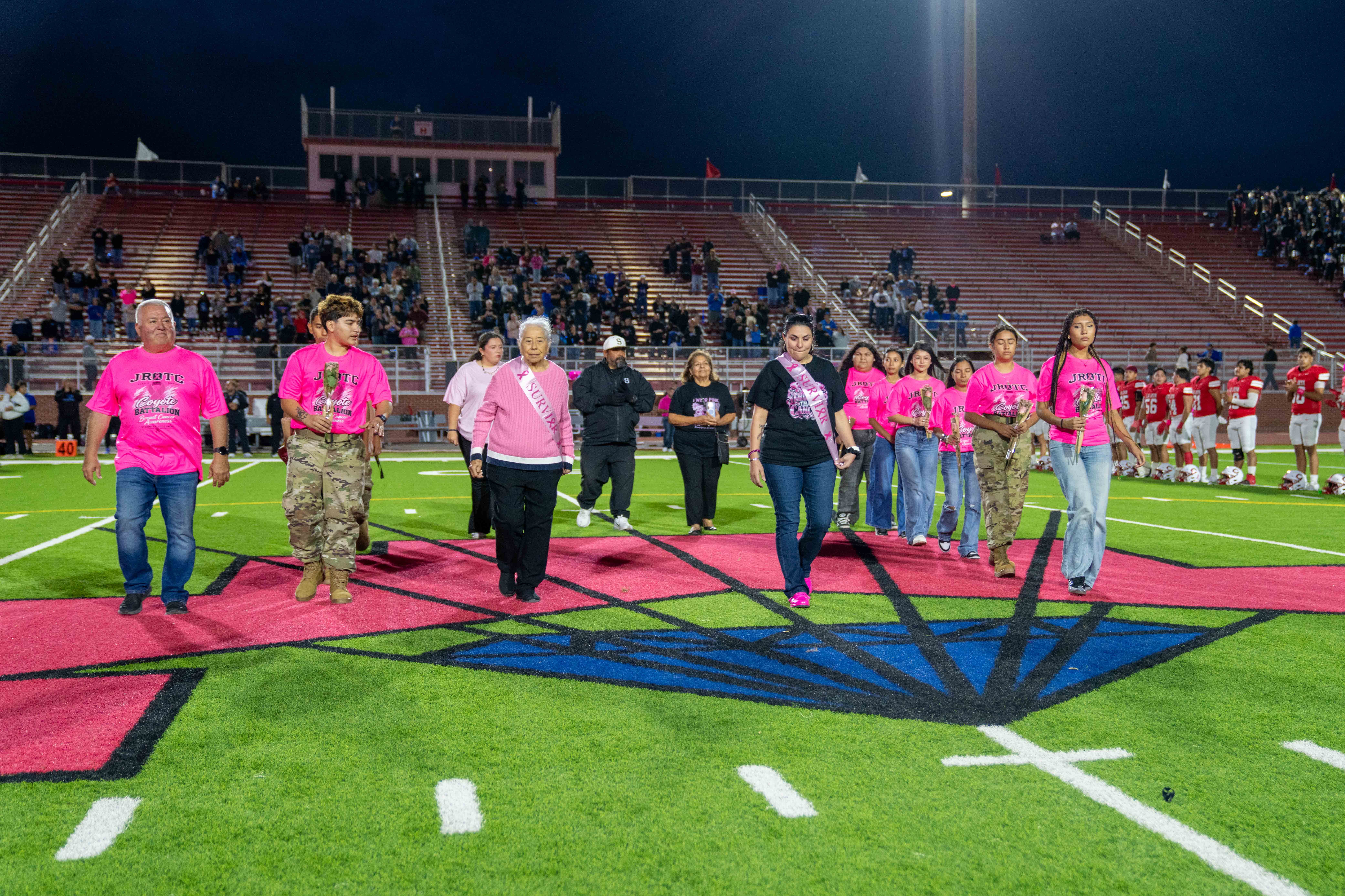 A group of people wearing pink shirts and sashes walk across a football field during a nighttime breast cancer awareness event.