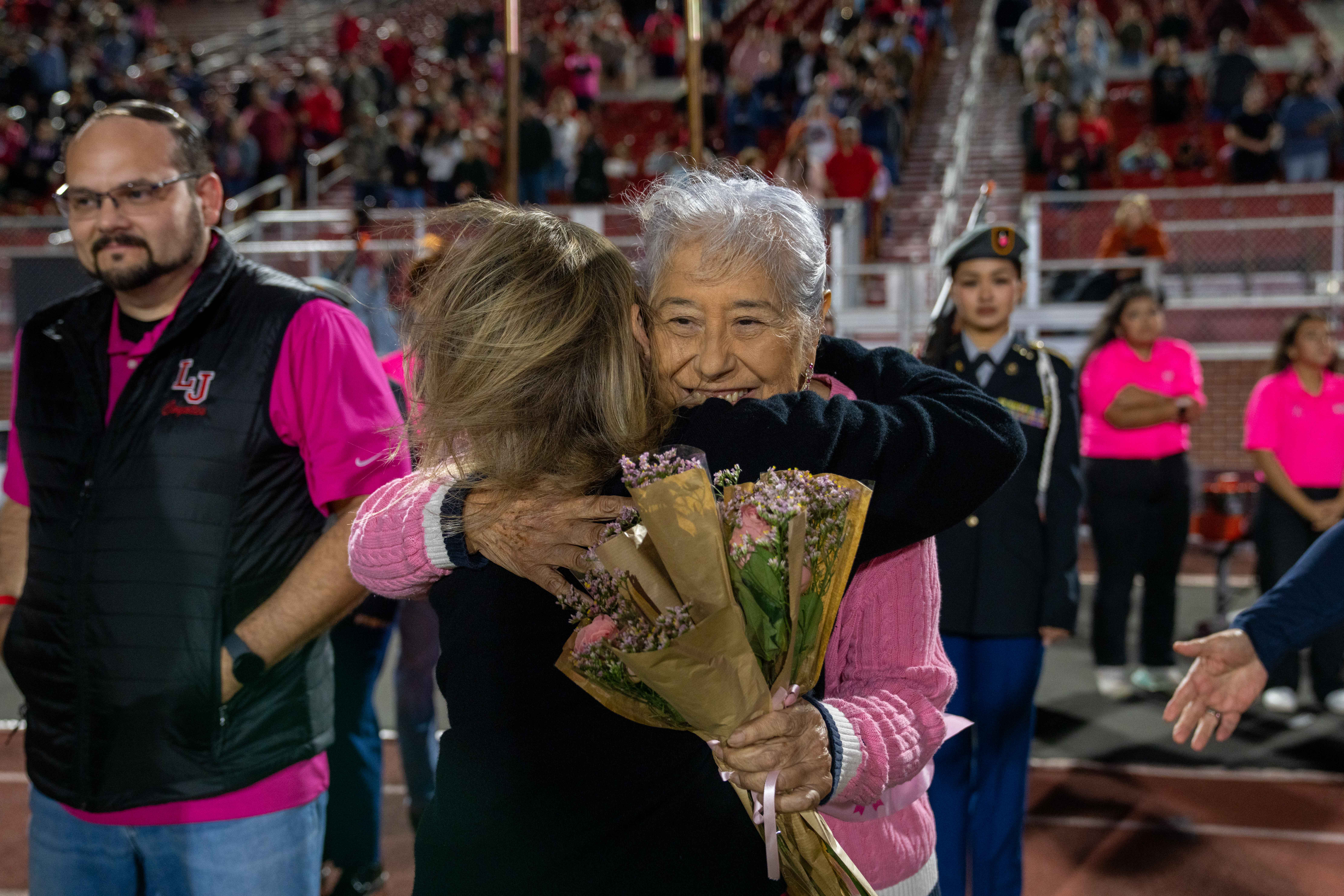 An older woman with silver hair smiles while embracing another person on a stadium track.