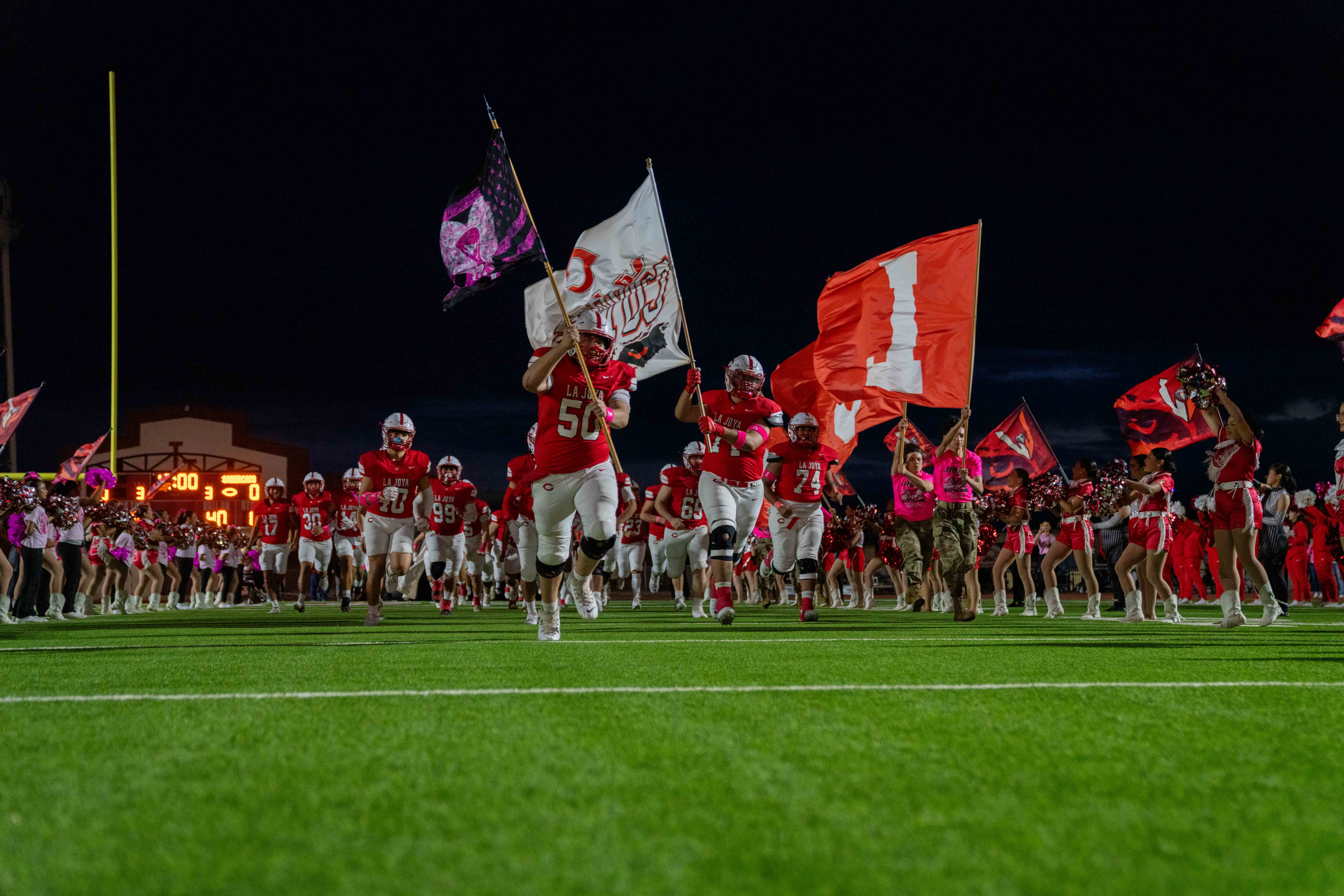 A high school football team runs onto the illuminated field at night, carrying flags.