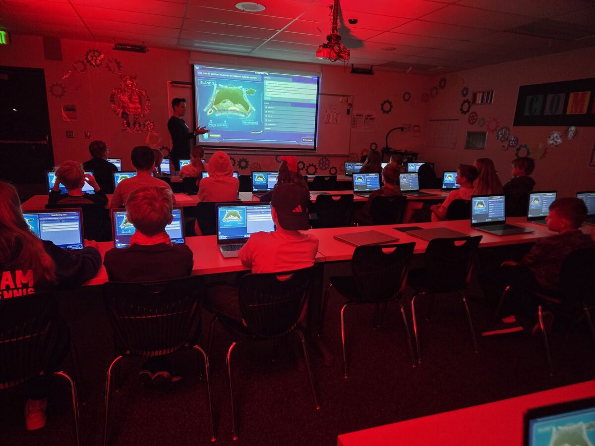 A presenter facilitates a Mission.IO lesson to students using laptops in a red-lit classroom.