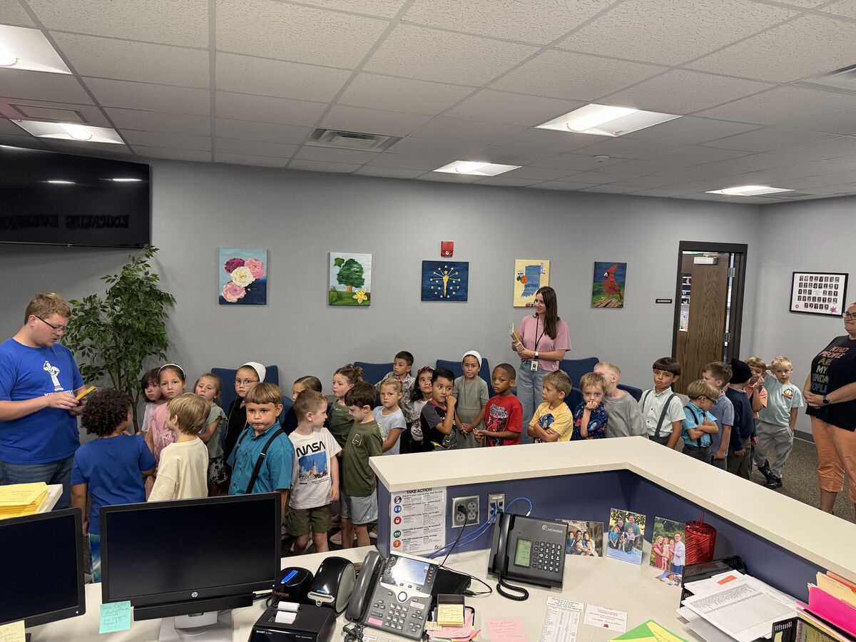A group of children stand in a line, listening to a man in a blue shirt and a woman in a pink shirt in an office setting.