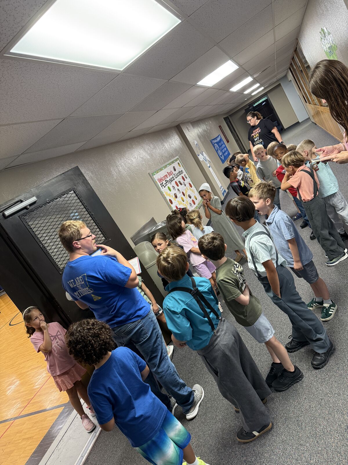 A group of students stand in a line indoors, some looking at a poster on the wall.