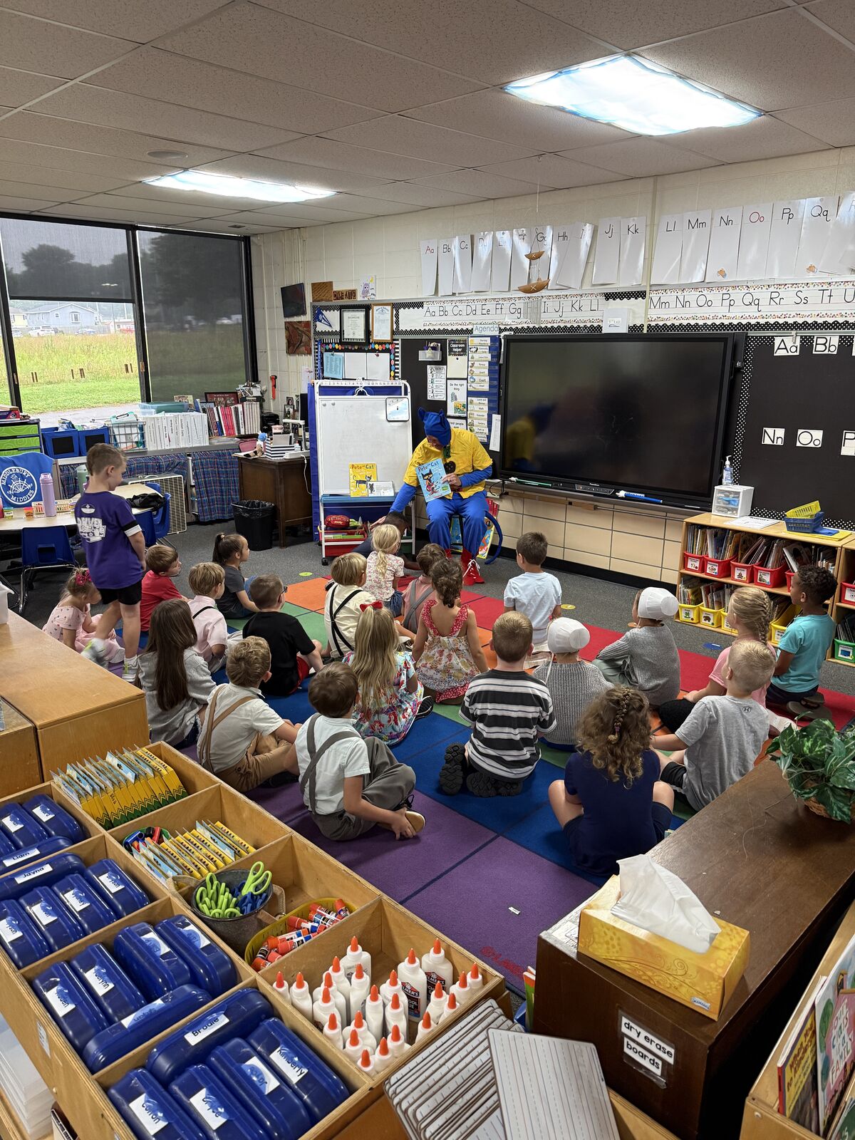 A teacher dressed as Pete the Cat reads a book to a group of young students sitting on the floor in a classroom.