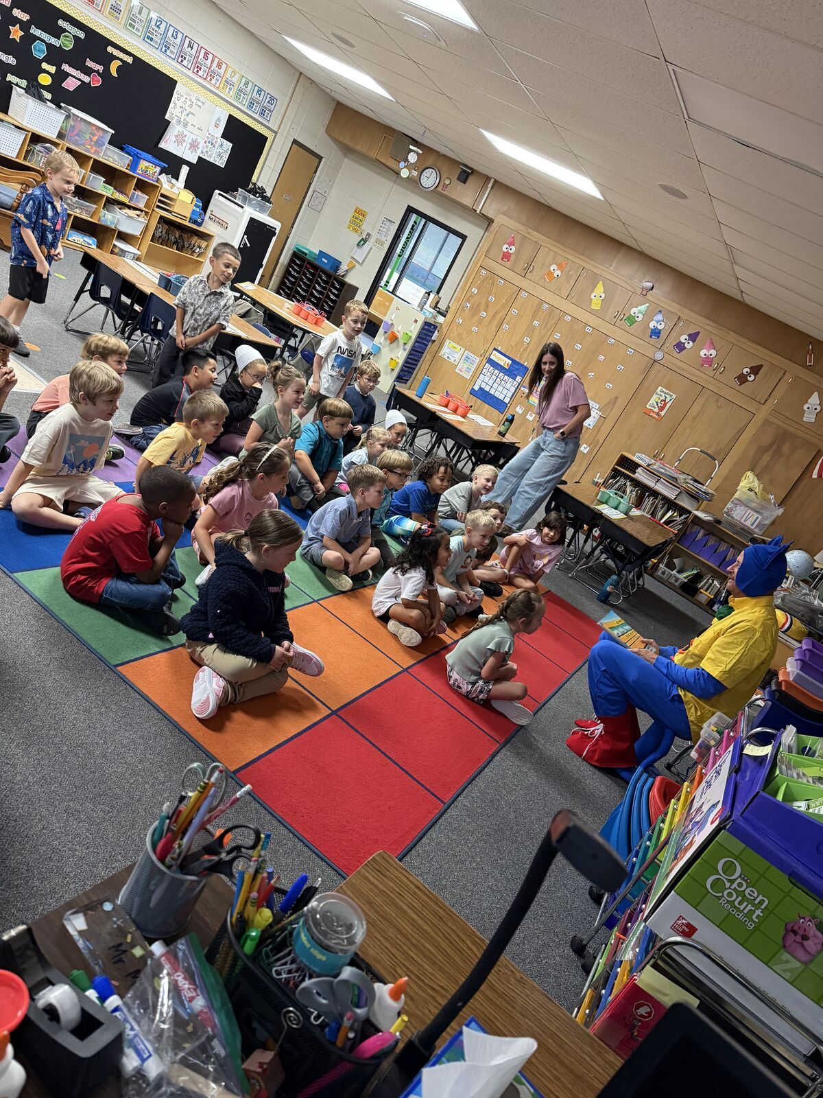 A group of young students sit on a colorful rug in a classroom, attentively watching a person in a blue and yellow costume.