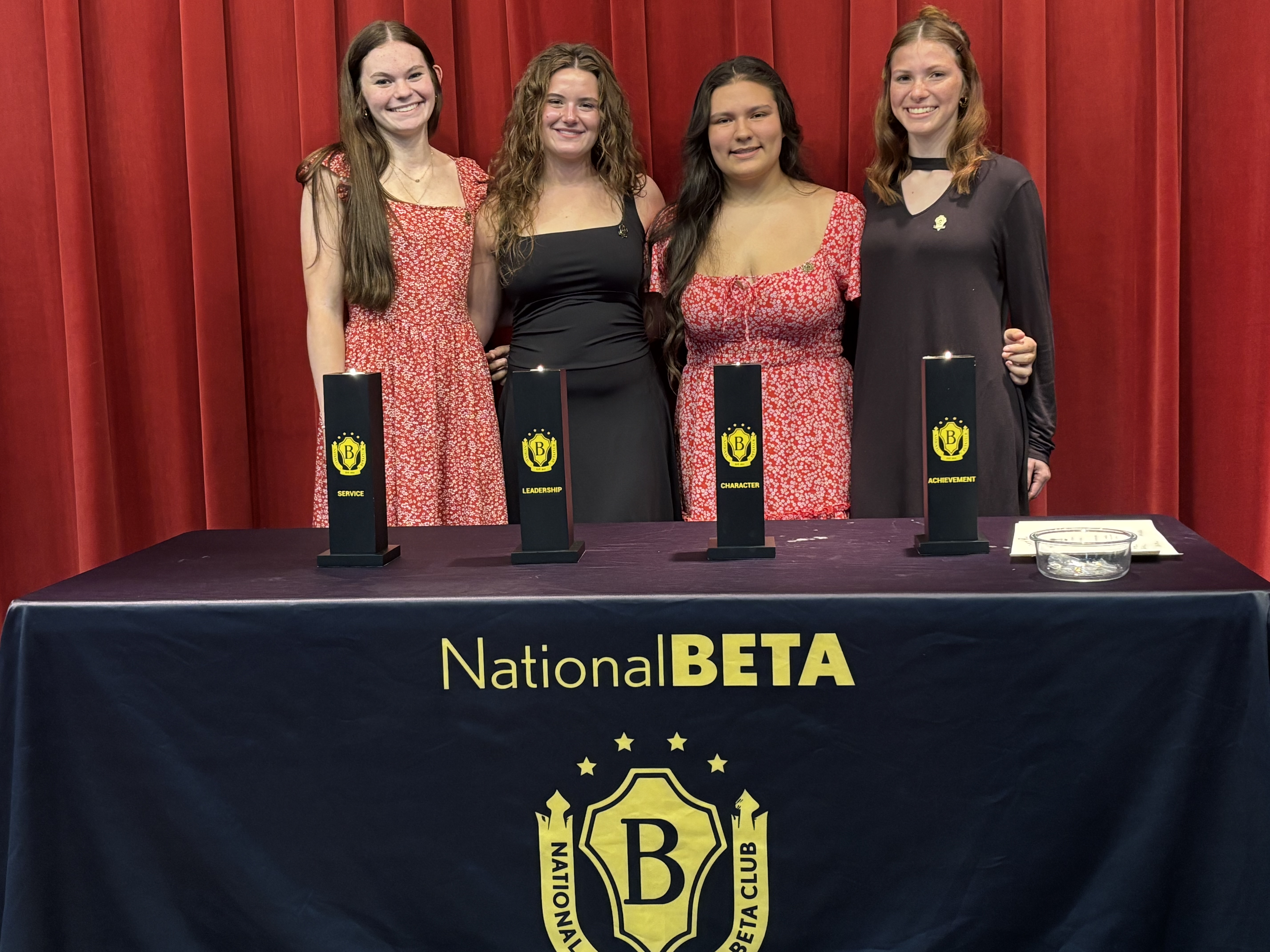 Four young women stand behind a table with a 'National BETA' cover, in front of a red curtain, with four pillars labeled Service, Leadership, Character, and Achievement.
