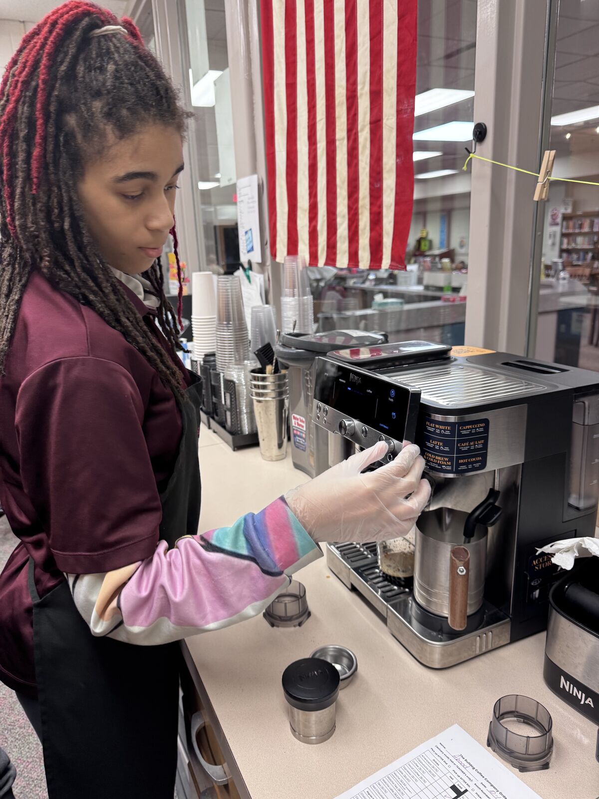 A barista with braided hair operates an espresso machine behind a counter, with an American flag hanging in the background.