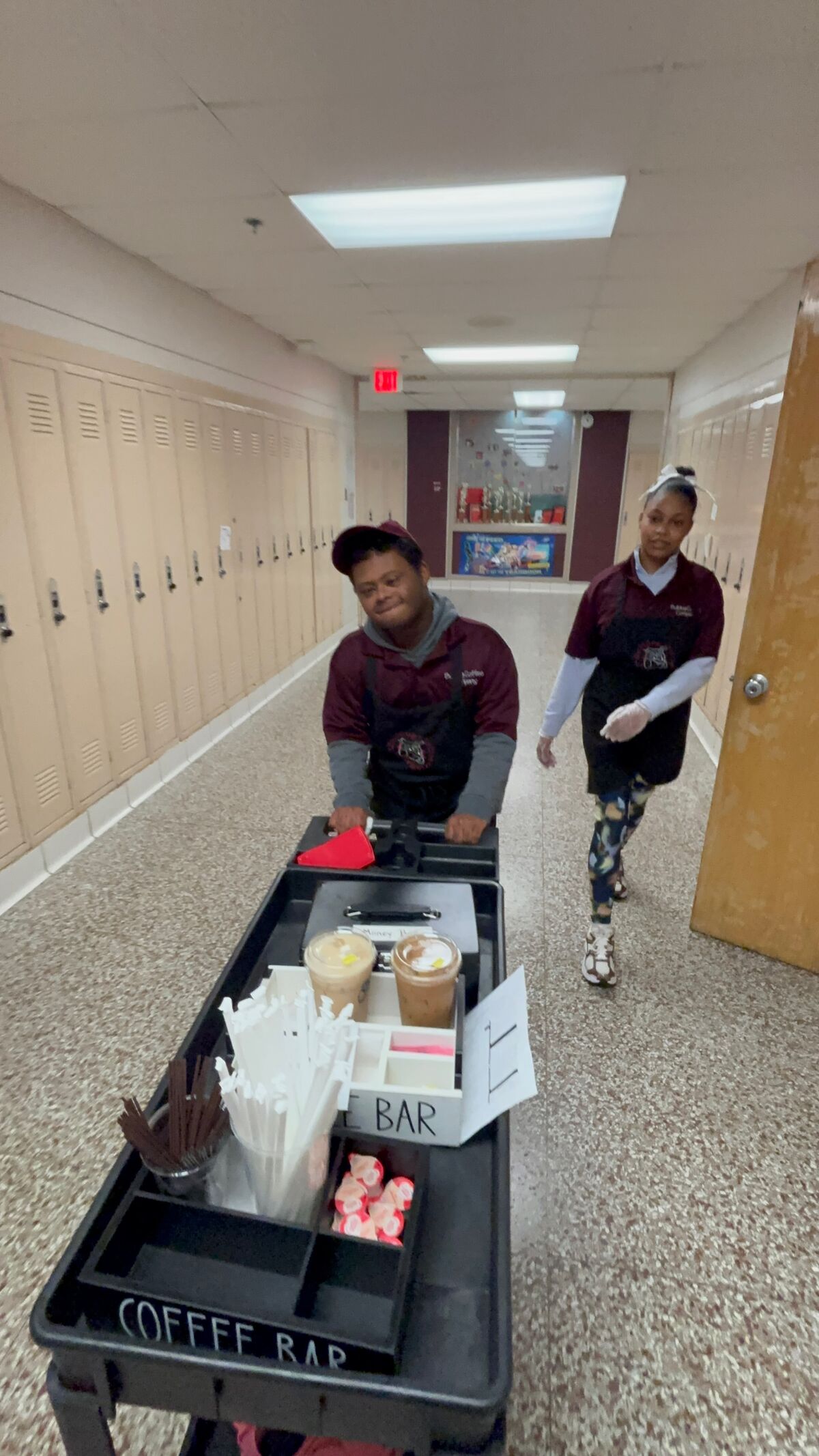 Two smiling students push a coffee cart stocked with drinks and supplies down a school hallway.