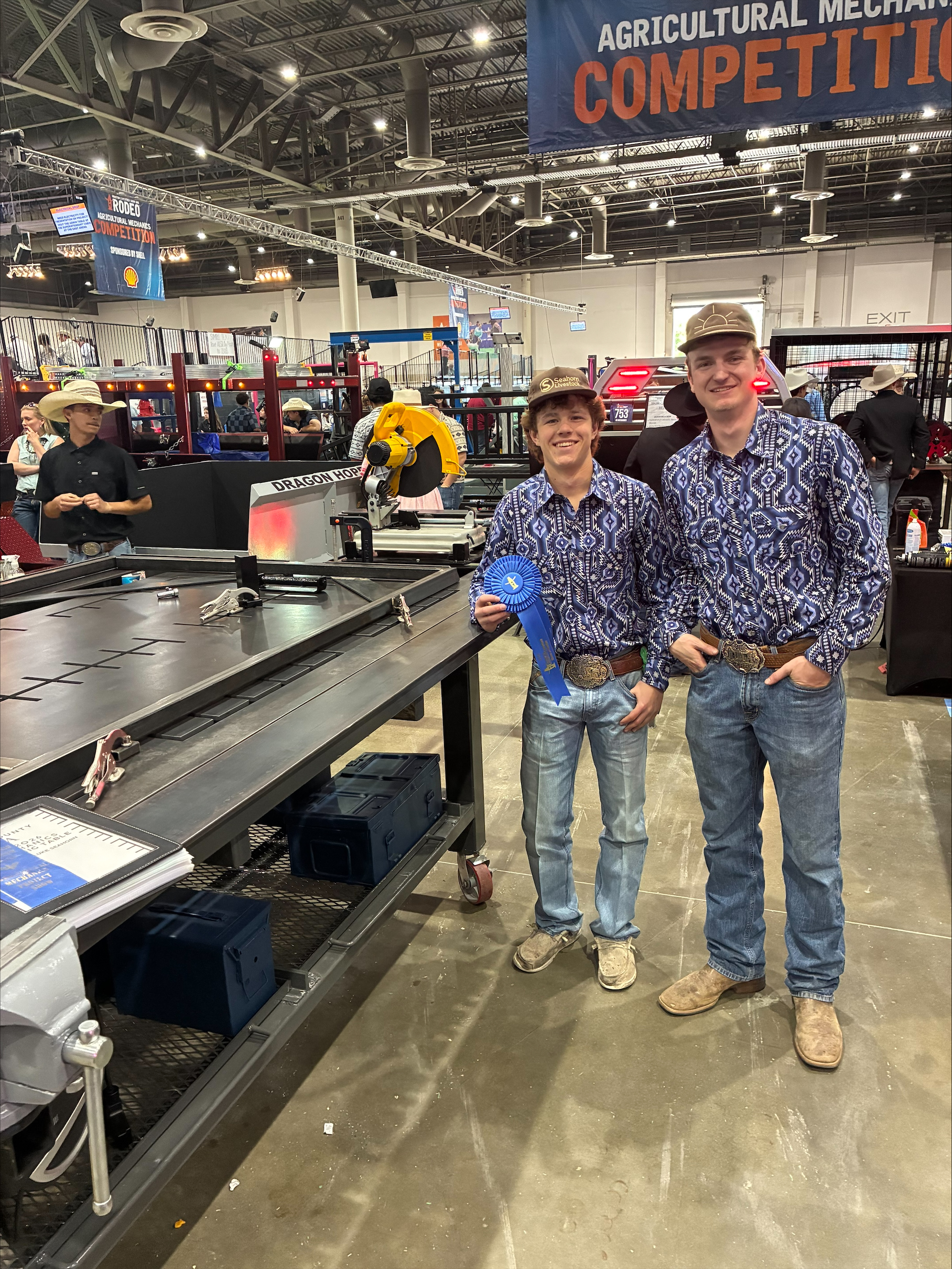 Two young men in western wear smile, one holding a blue ribbon, at an agricultural mechanics competition.