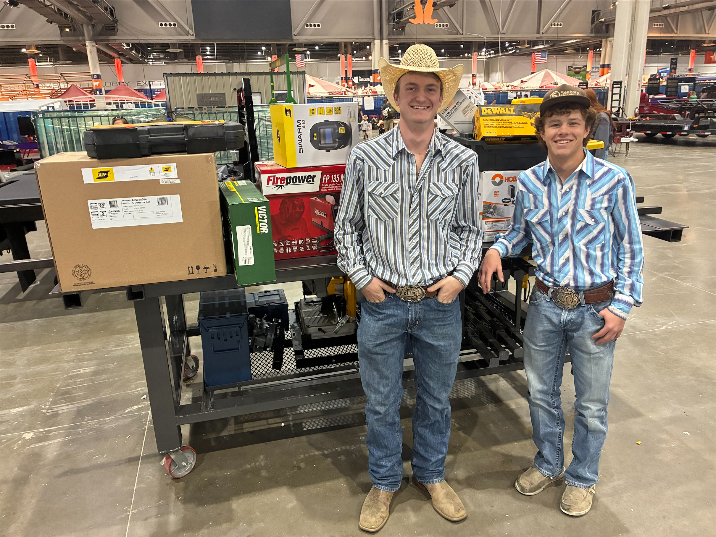 Two young men in western shirts and jeans stand next to a table displaying various tools and equipment, including boxes from ESAB, Firepower, and DeWalt.