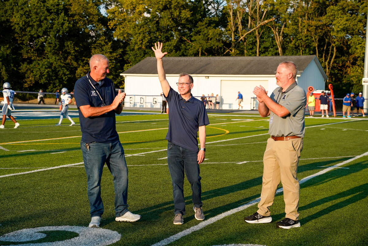 Three men stand on a football field, two clapping and one waving, with a building and trees in the background.