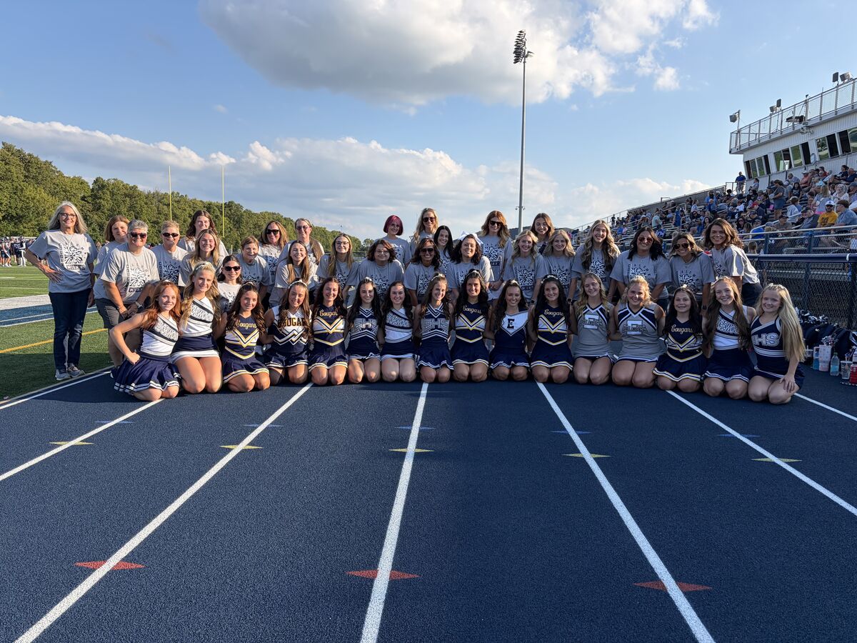 A large group of cheerleaders and adults pose for a photo on a blue running track with stadium seating in the background.