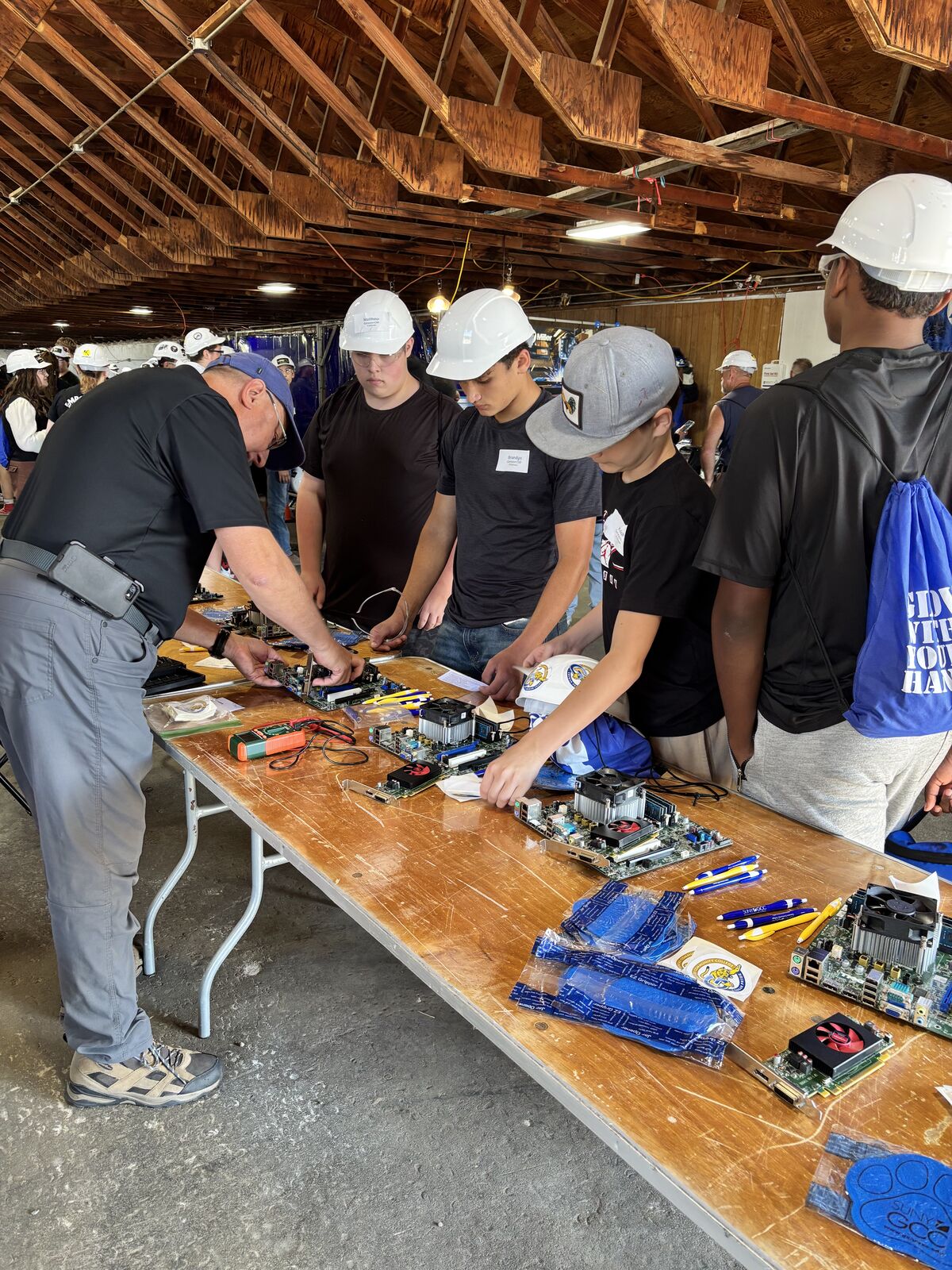 People gather around a long wooden table examining computer hardware components and circuit boards.