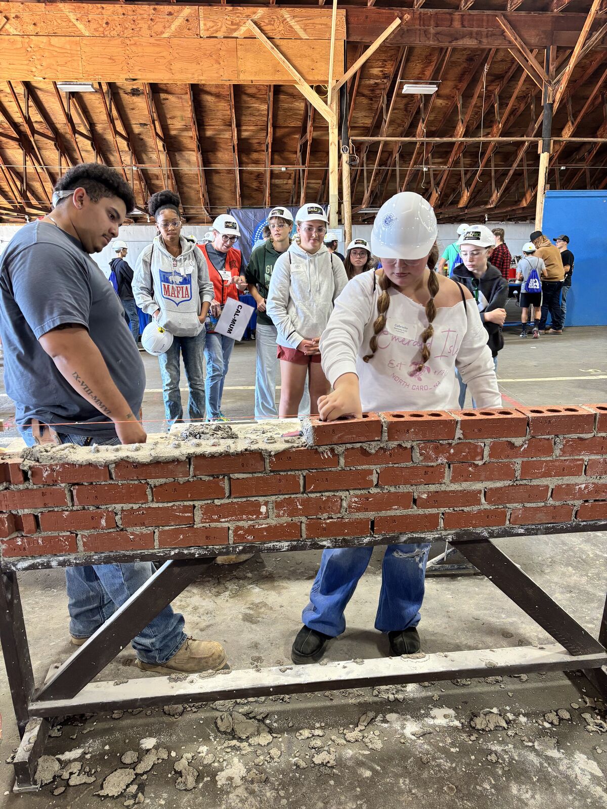 A person wearing a white hard hat carefully places a red brick on a wall inside a large workshop.