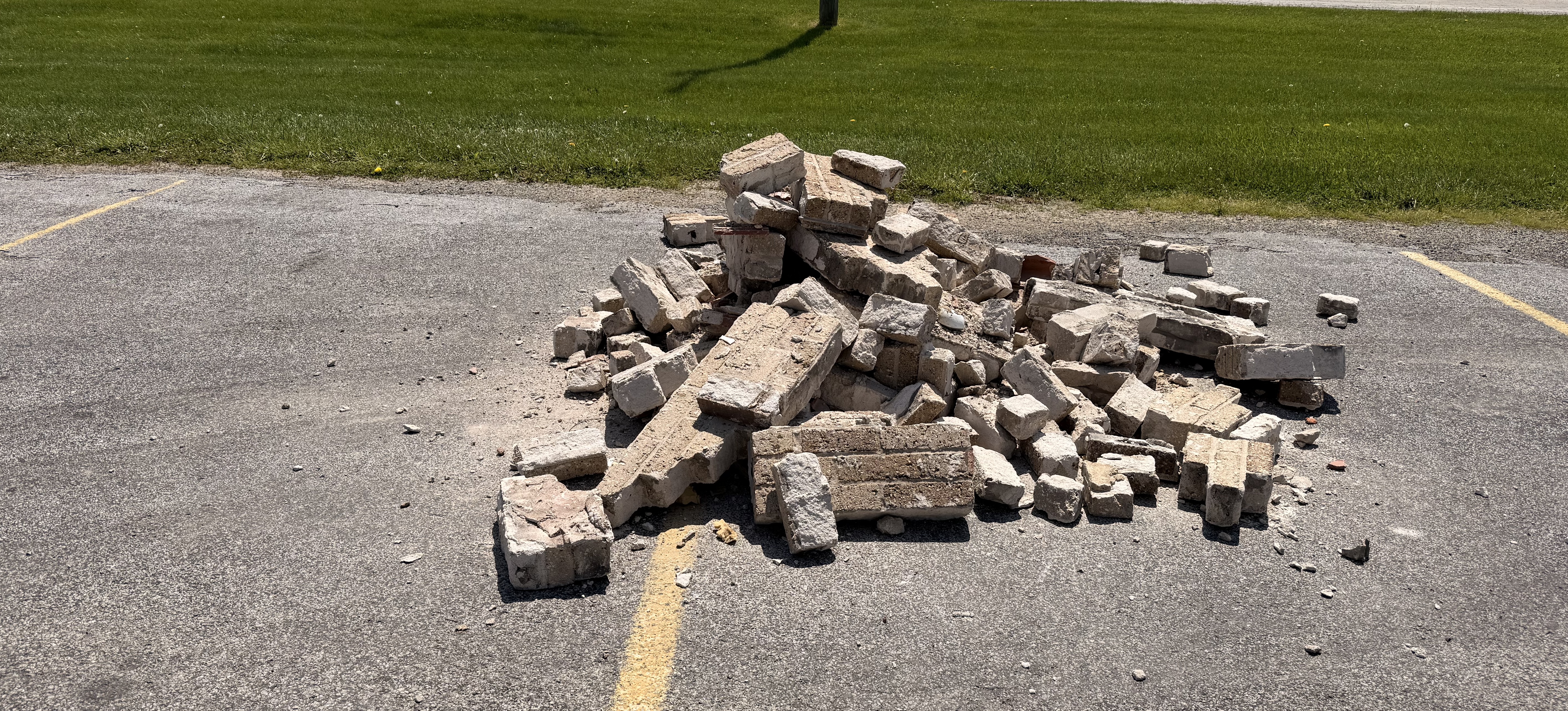 A pile of broken bricks lies on an asphalt surface next to a grassy area.