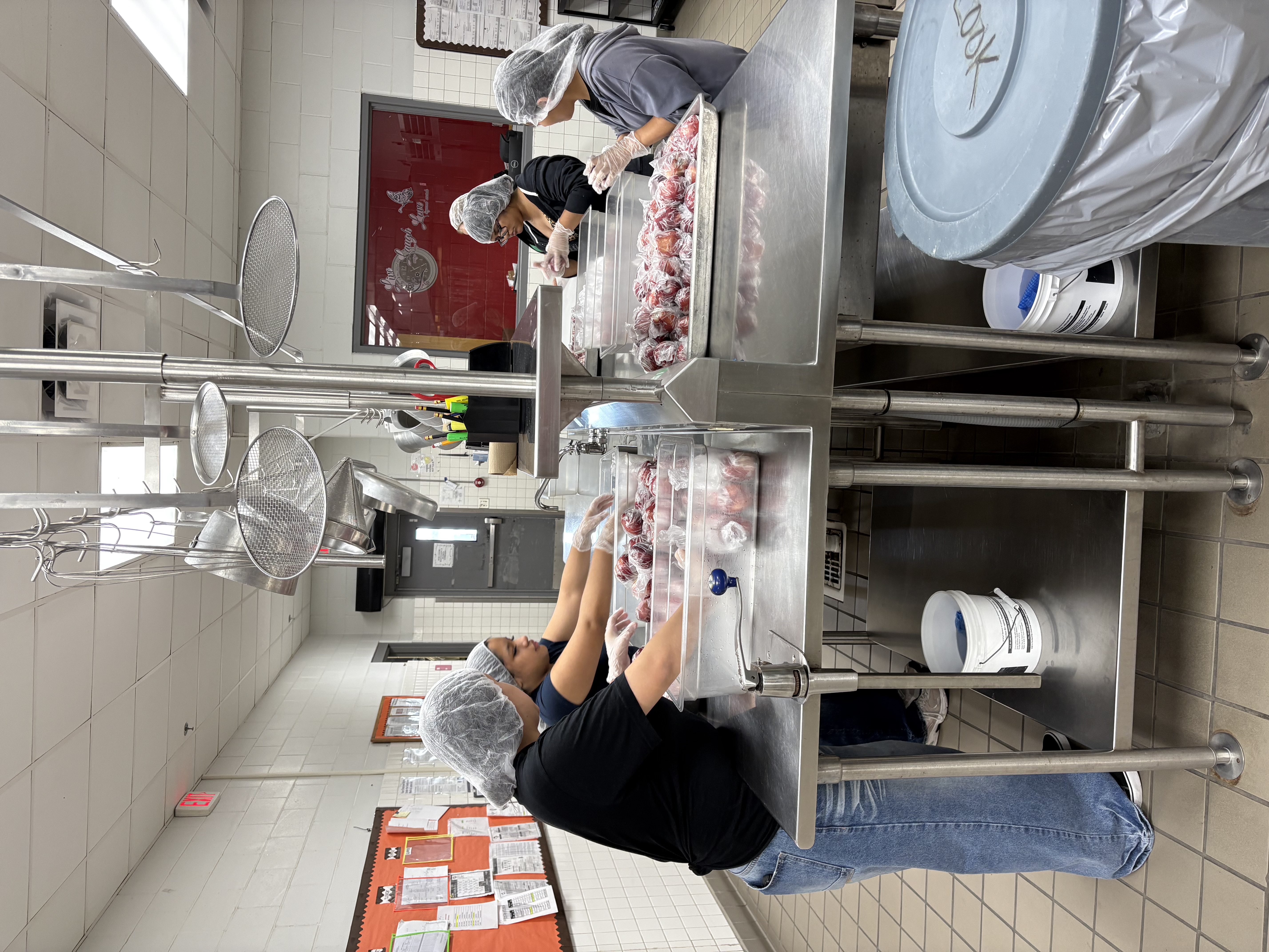 Three individuals wearing hairnets and gloves prepare food items in a commercial kitchen setting.