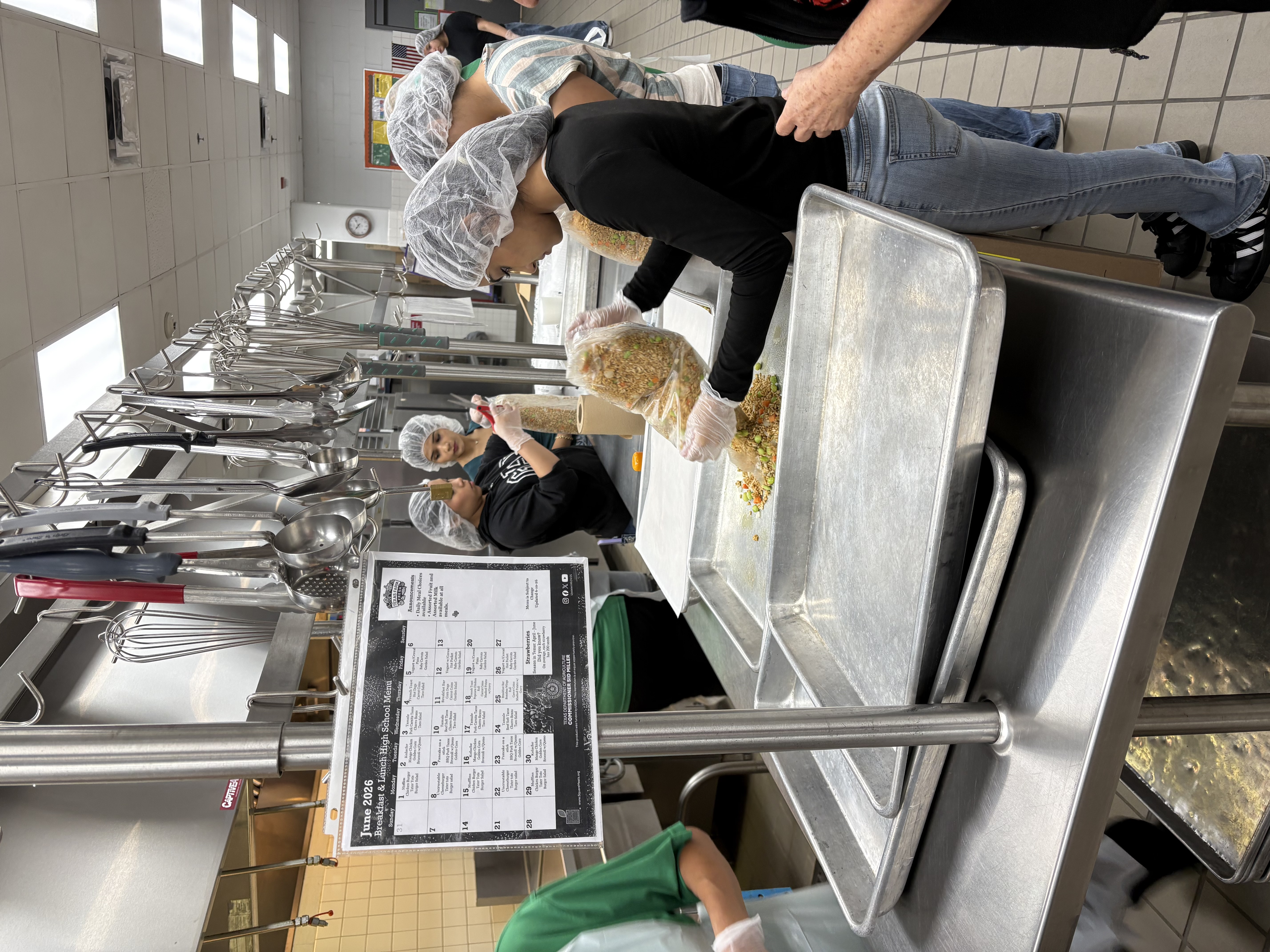 People in hairnets and gloves prepare food in a commercial kitchen, pouring ingredients from plastic bags into large metal trays.
