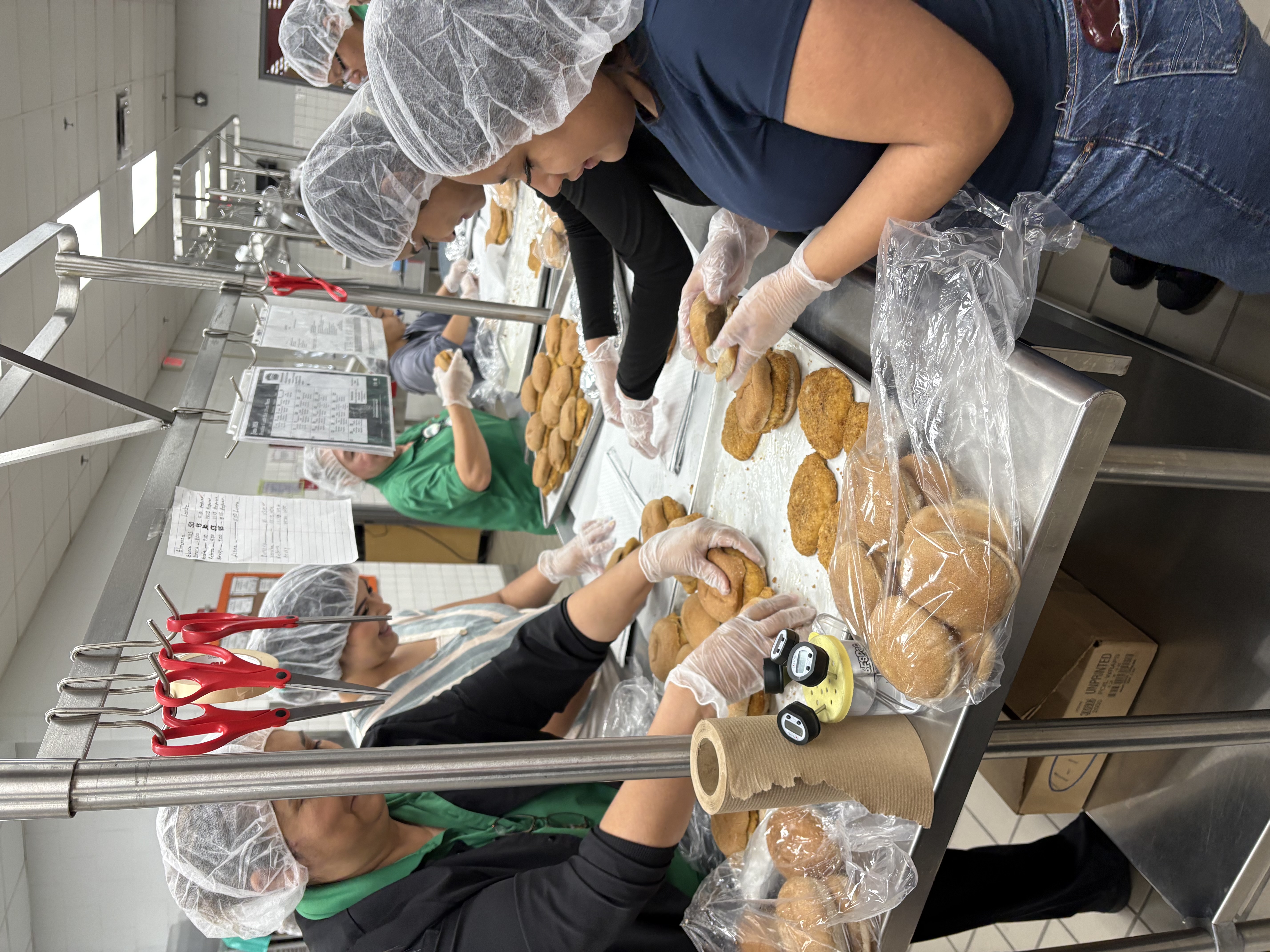 People in hairnets and gloves assemble food items on a stainless steel counter.