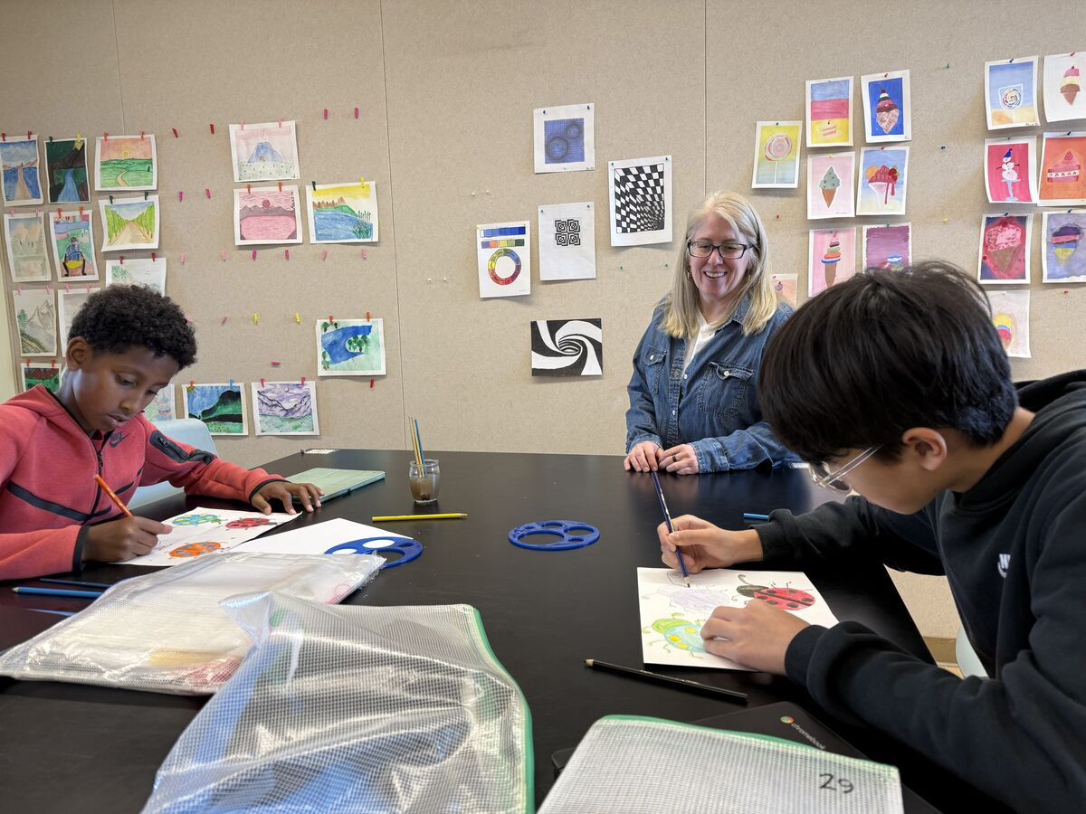 A teacher smiles as two students draw ladybugs at a table in an art classroom.