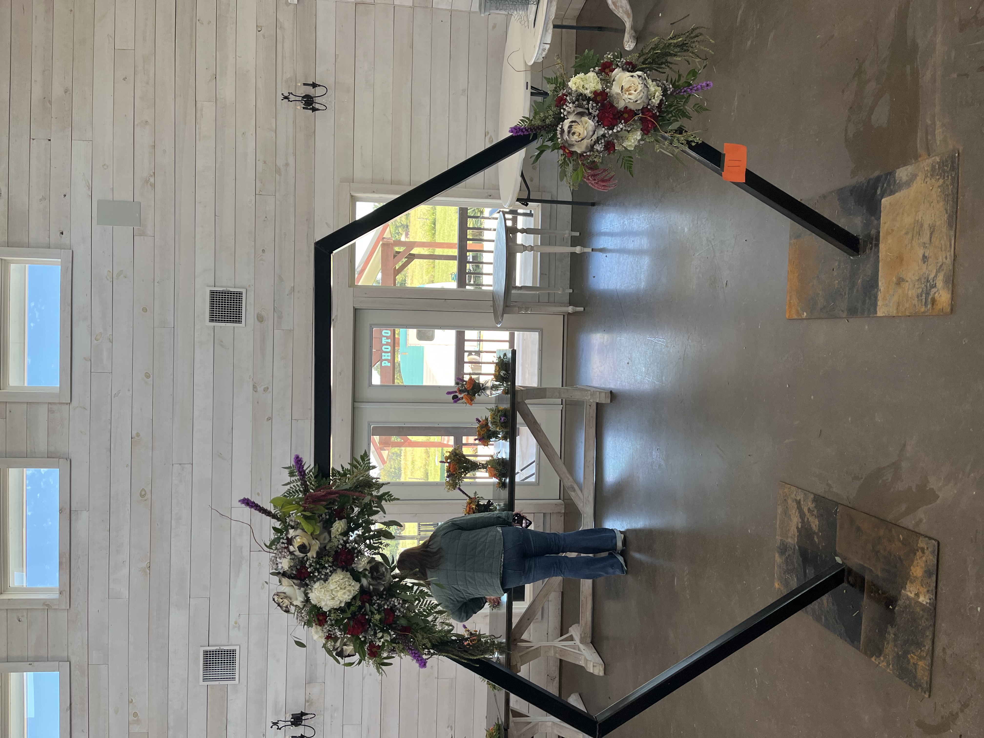 A person stands inside a rustic room with white wooden walls, preparing a black geometric wedding arch decorated with large floral arrangements.