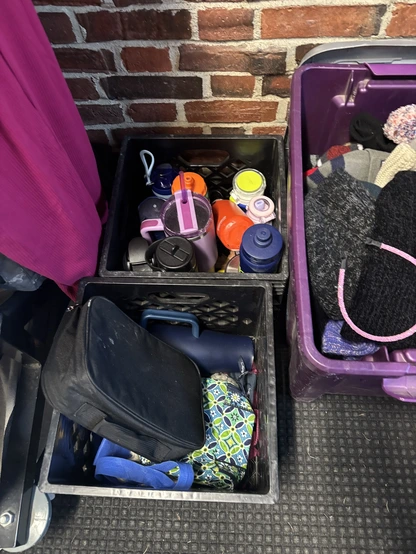 Several reusable water bottles and containers are neatly organized in black crates against a brick wall.