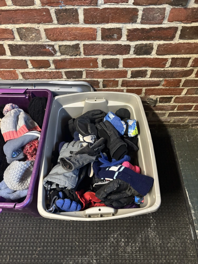 Two storage bins filled with winter hats and gloves are placed against a brick wall.