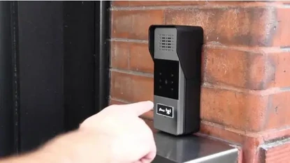 A finger presses a button on a modern intercom system mounted on a brick wall.