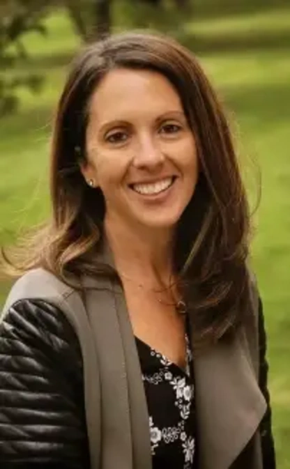 A smiling woman with brown hair and a patterned shirt looks directly at the camera.