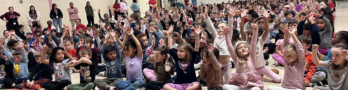 A large group of children and adults are seated on the floor, with many raising their hands for PAWS UP.