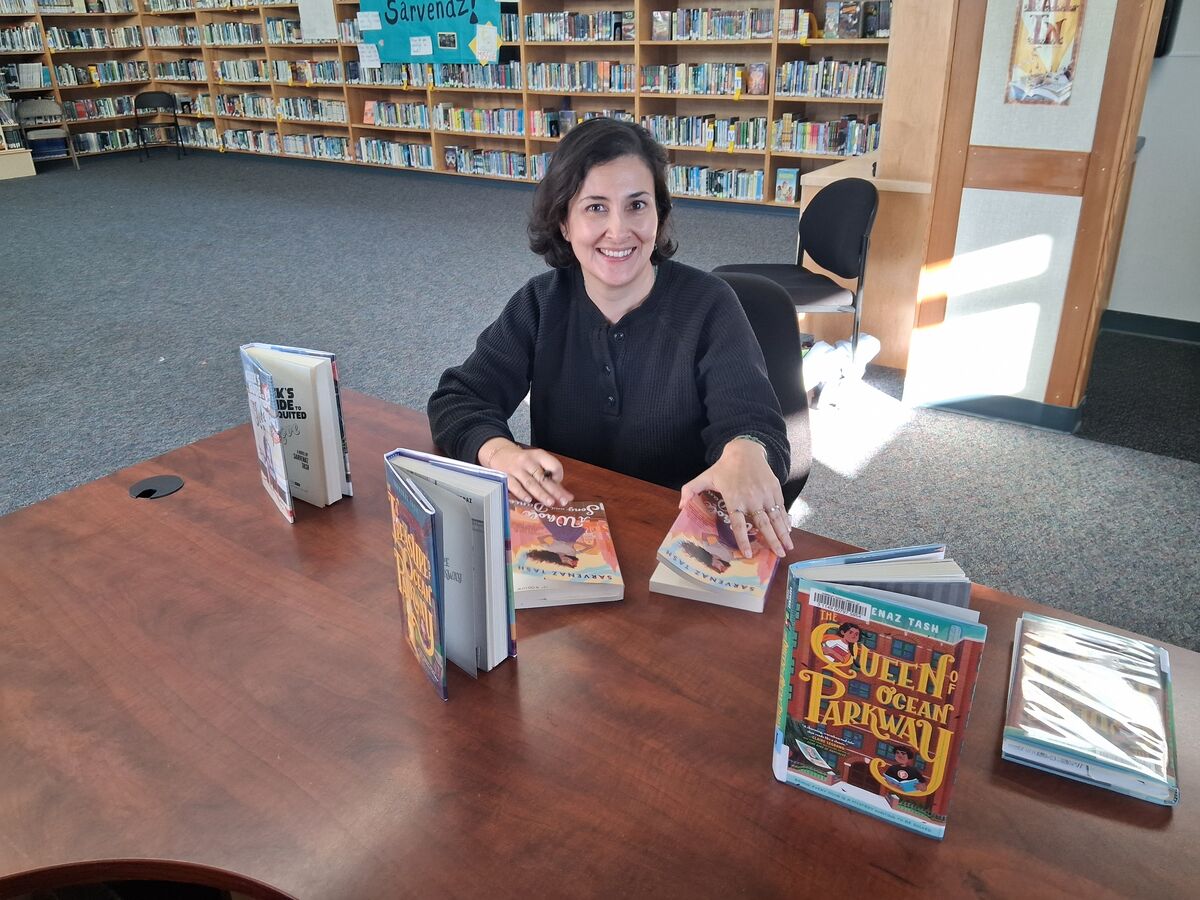 A smiling author sits at a wooden table in a library, surrounded by several copies of her book.