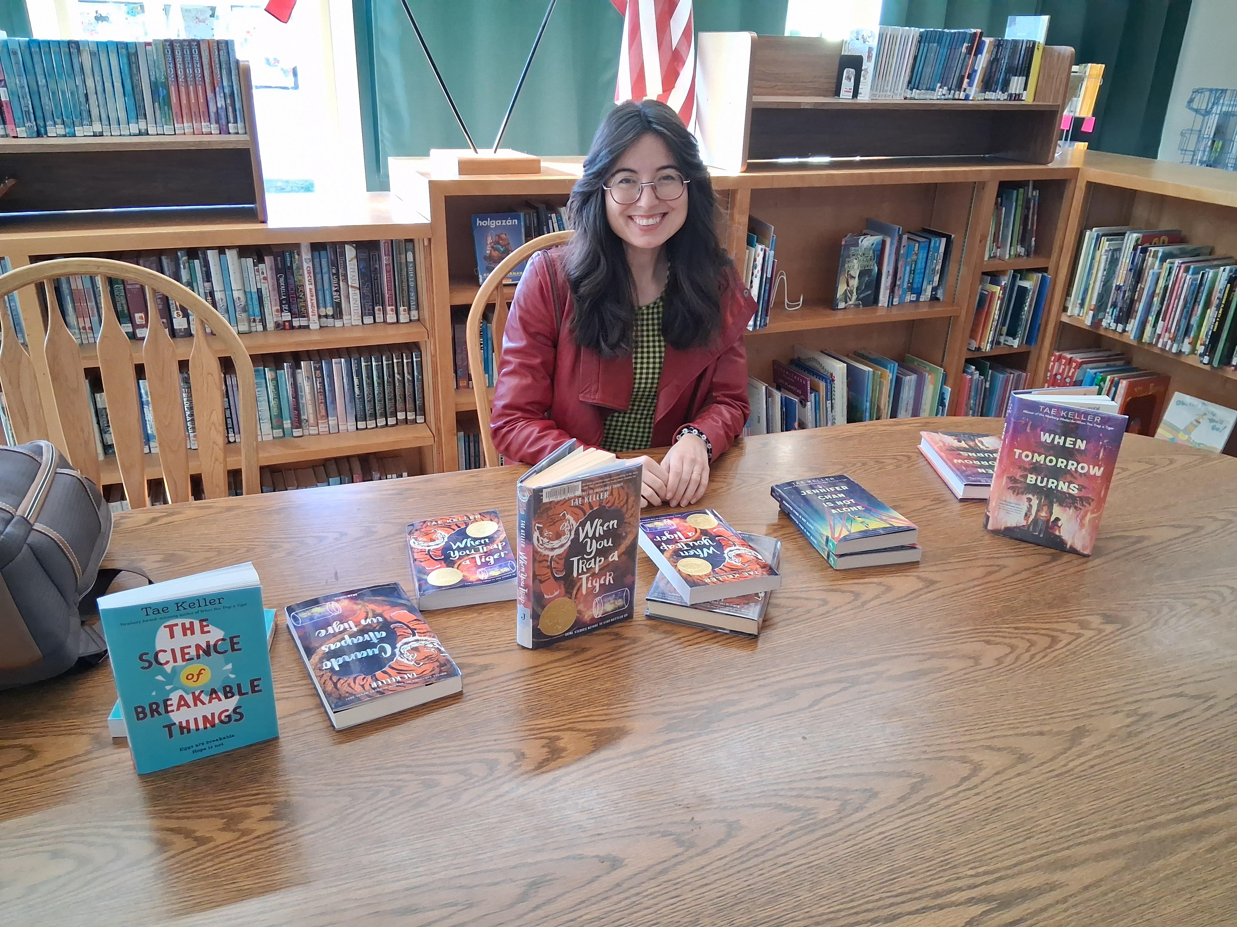 A smiling author sits at a wooden table in a library with copies of her books displayed.