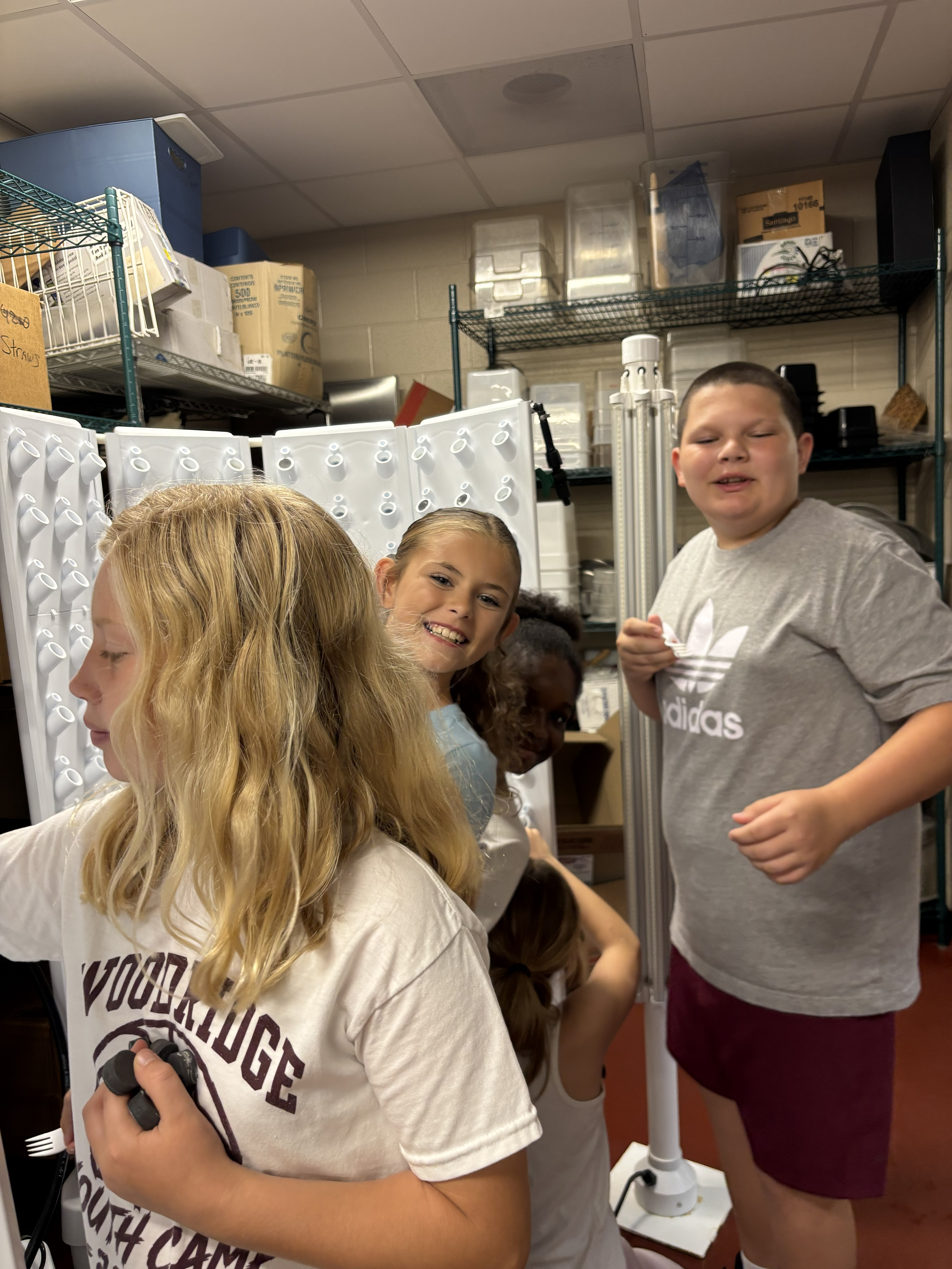 Four children smile and interact in a storage room filled with shelves and white equipment.