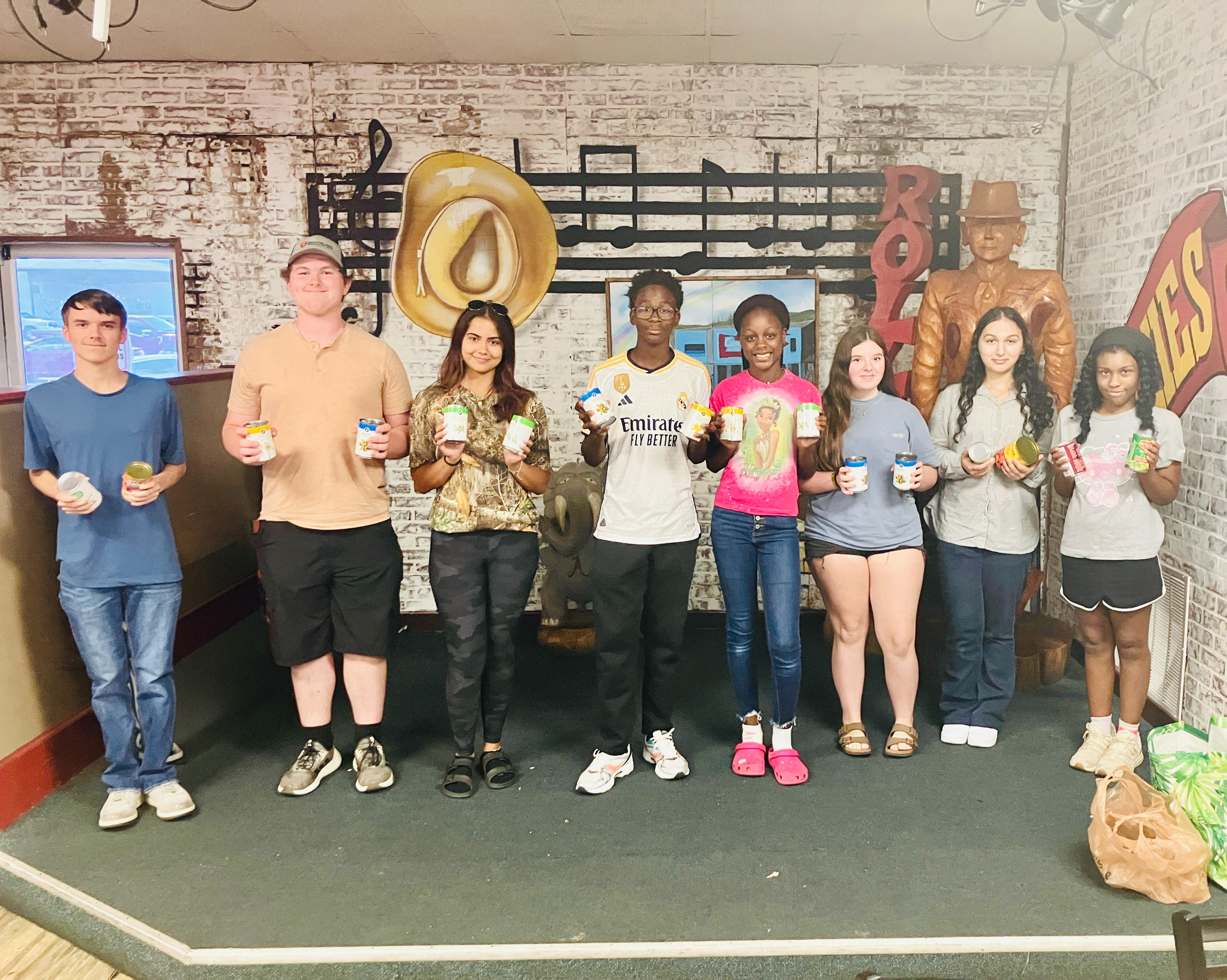 A group of eight diverse young people stand in a line, smiling and holding canned goods in front of a music-themed brick wall.