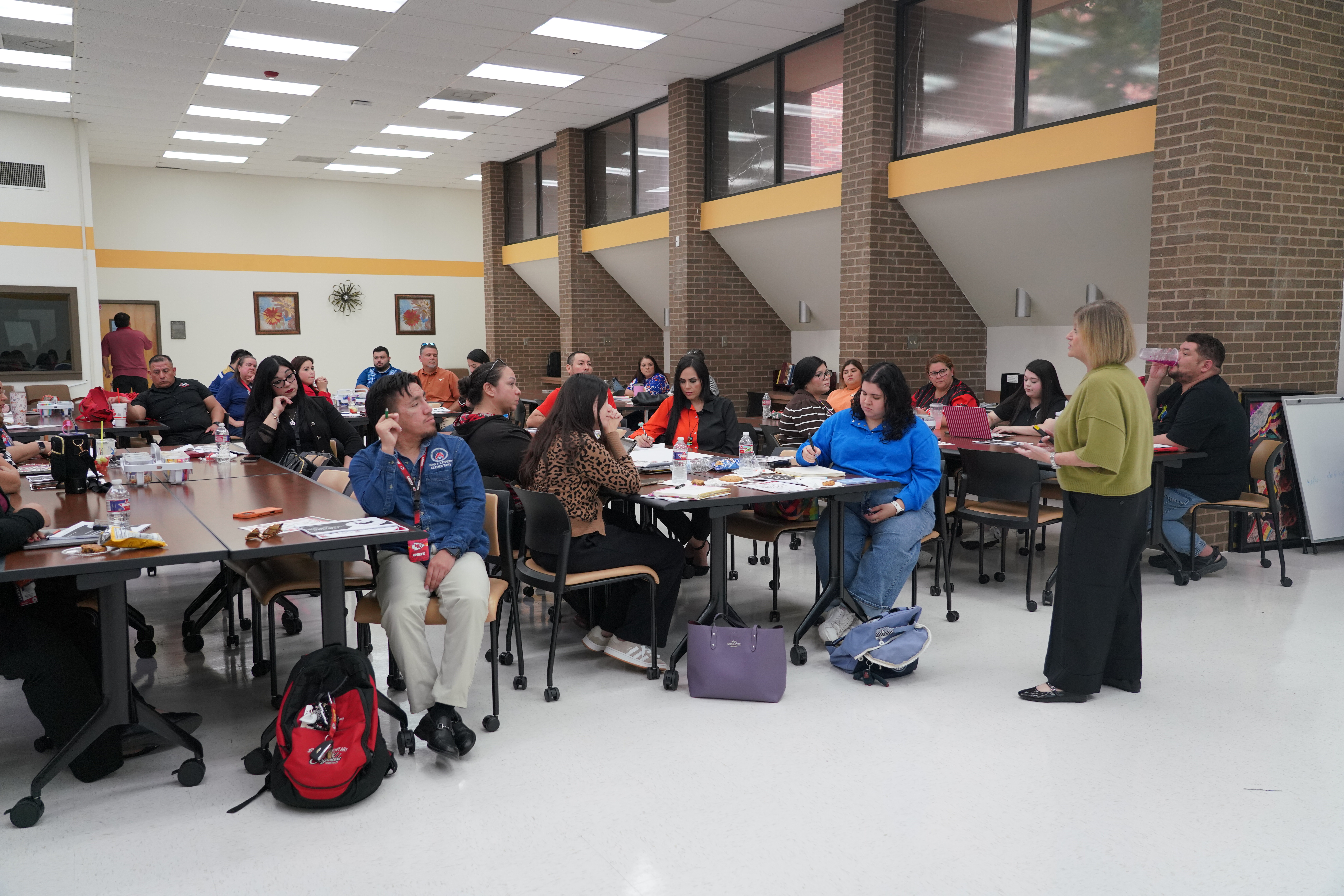 A group of adults sits at tables in a bright, brick-walled room while a woman stands and speaks.