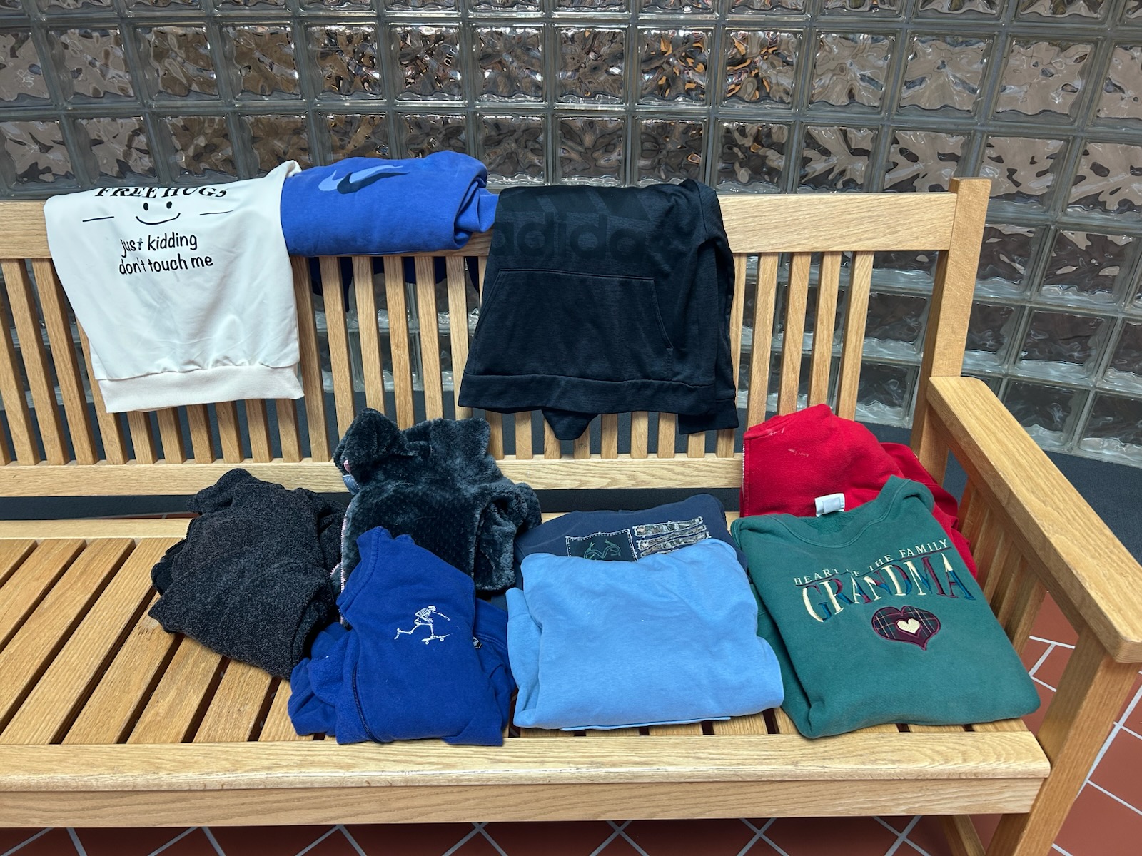 A wooden bench indoors holds several folded hoodies and sweatshirts of different colors.