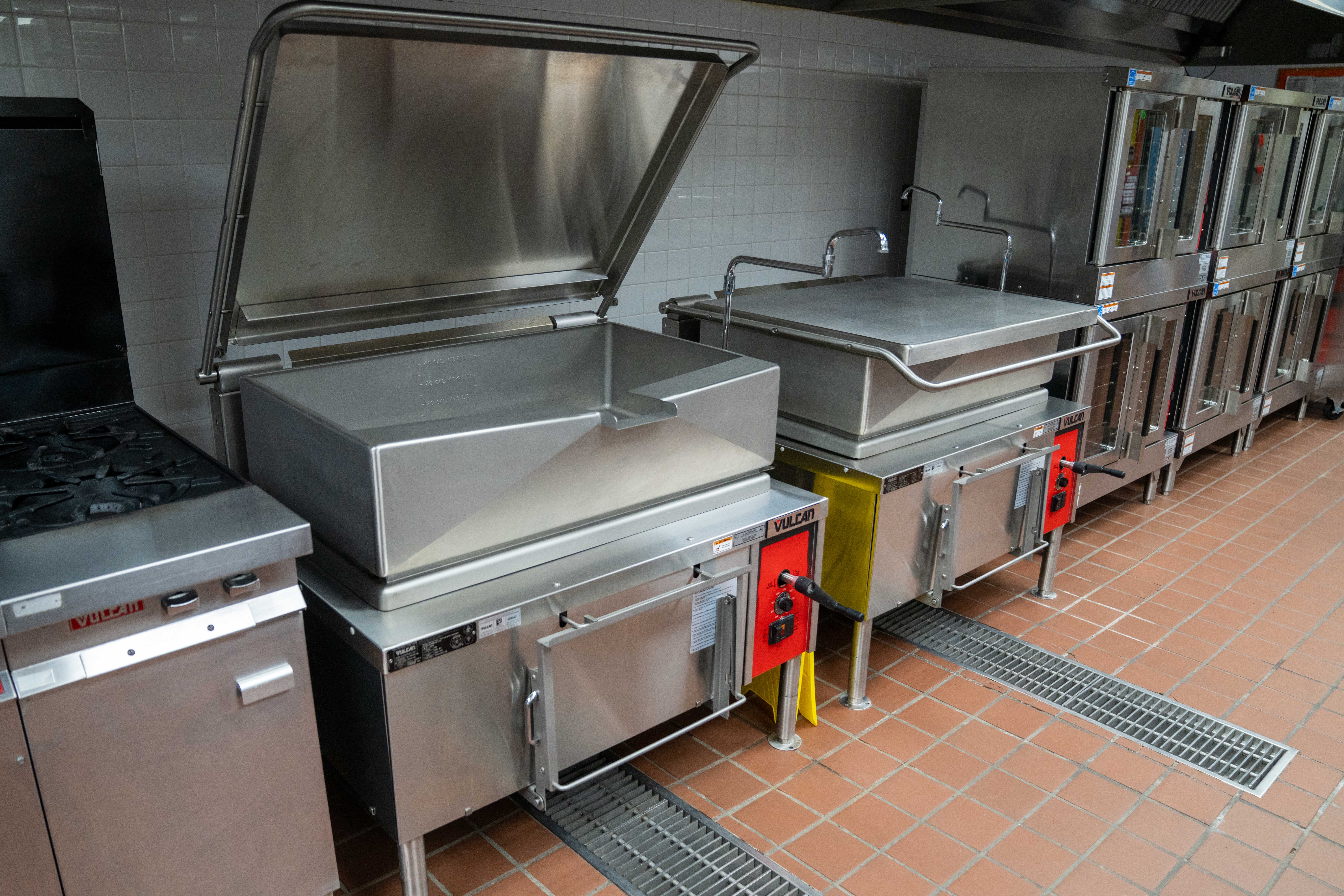 Commercial kitchen with stainless steel cooking equipment, including a large open kettle and stacked ovens.