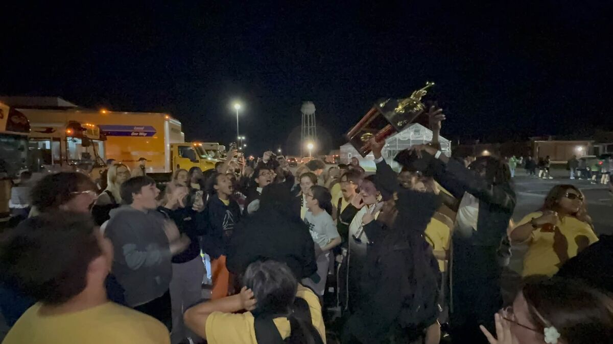A crowd of people cheers enthusiastically at night, holding up a large trophy in celebration.