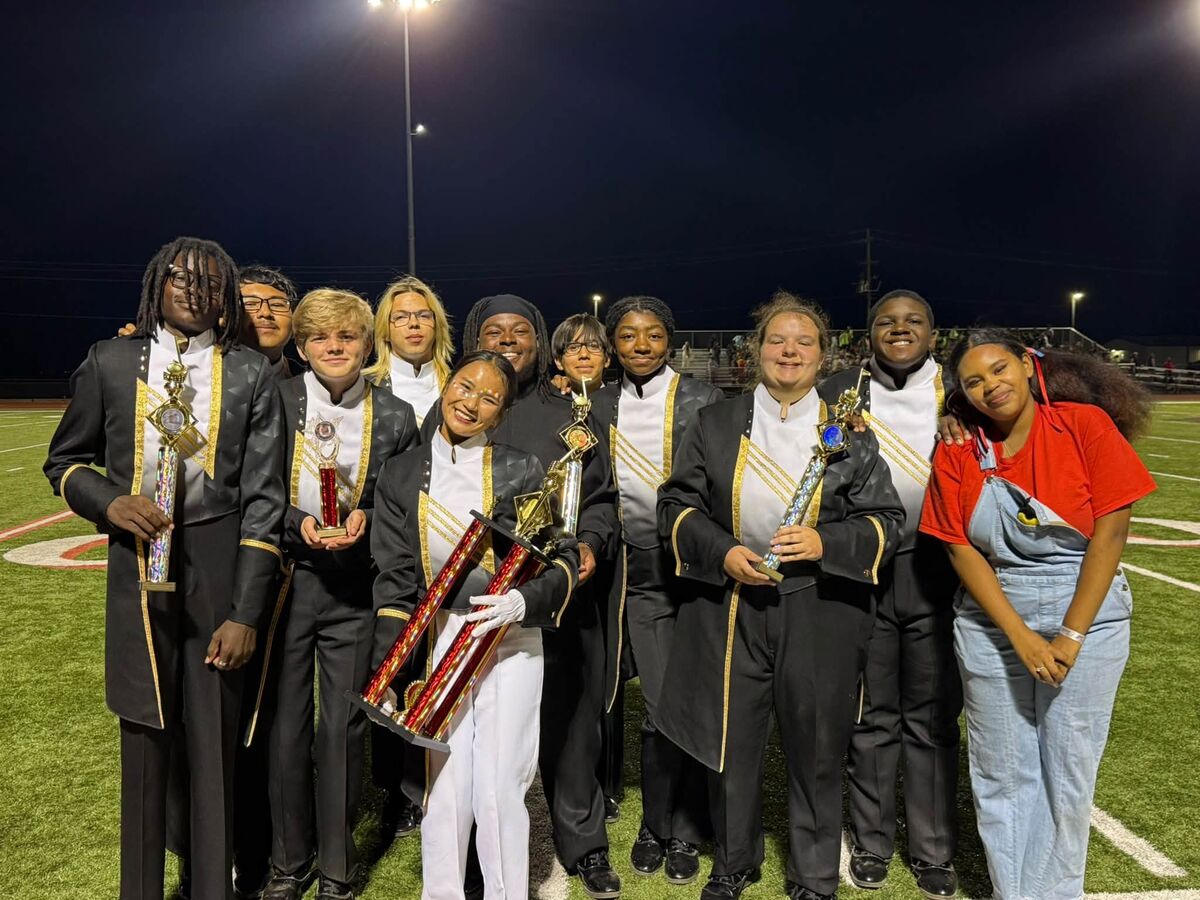 A group of smiling marching band members proudly hold trophies on a lighted football field at night.