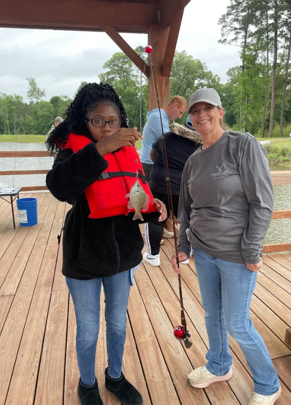A young person in a red life vest proudly holds a small fish while fishing with an adult on a wooden dock.