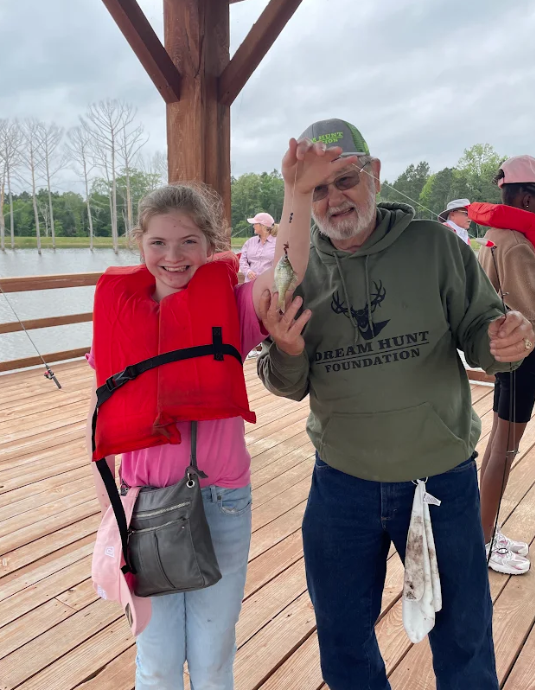 A smiling girl in a red life jacket stands next to a man holding a small fish.