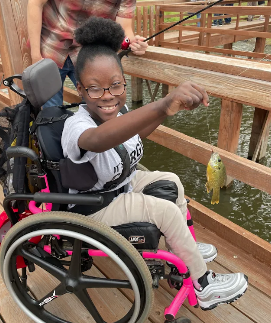 A young person in a wheelchair proudly holds up a small fish they just caught while fishing from a wooden pier.