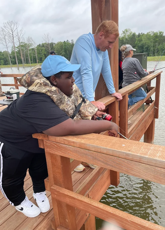 A young person in a life vest and blue cap fishes from a wooden pier, with an adult standing nearby.