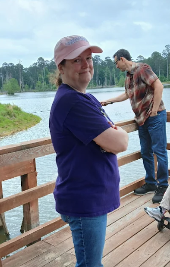 A woman in a pink cap and purple shirt stands on a wooden pier, smiling at the camera.