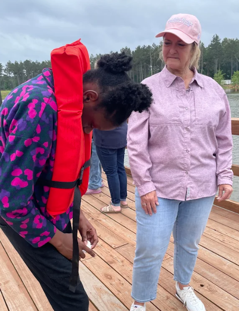 A young person wearing a bright orange life vest bends over on a wooden dock.