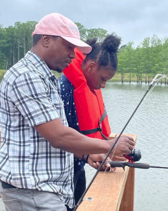 A man and a child fish together on a wooden dock by a lake.